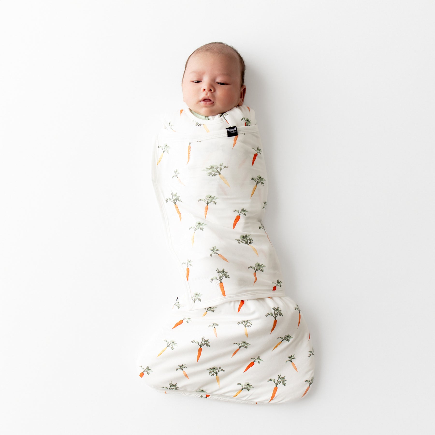 Newborn baby swaddled in the Sleep Bag Swaddler in Carrot 1.0 laying on a white surface