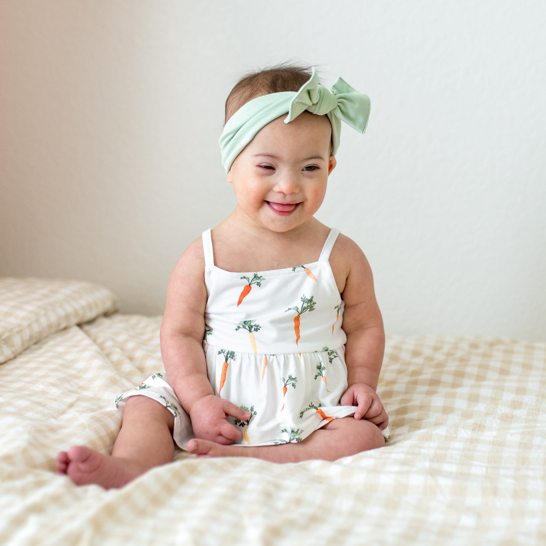 Young toddler sitting on a bed wearing the Spaghetti Strap Ruffle Romper in Carrot with a light green bow