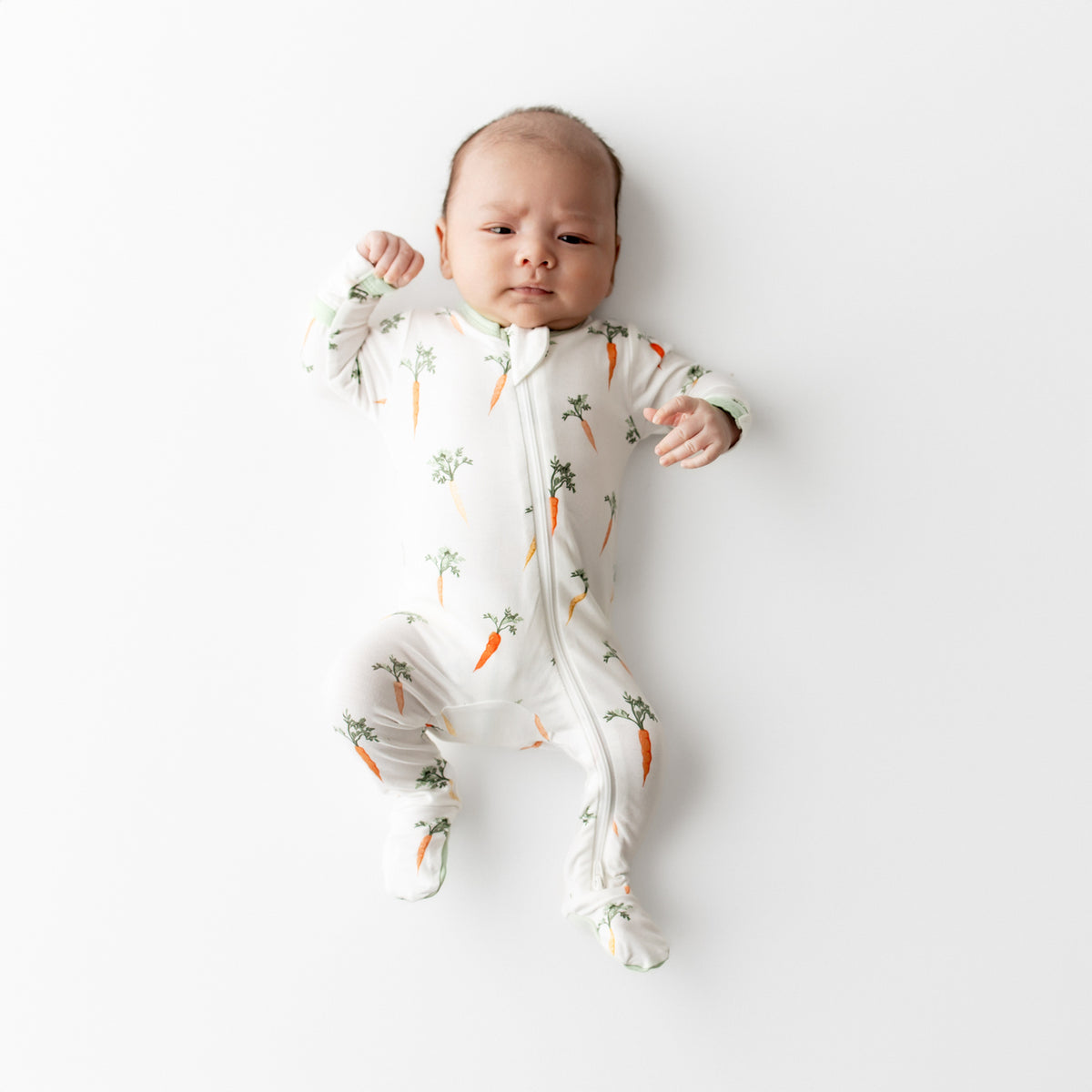 Newborn laying on a white surface wearing the Zippered Footie in Carrot