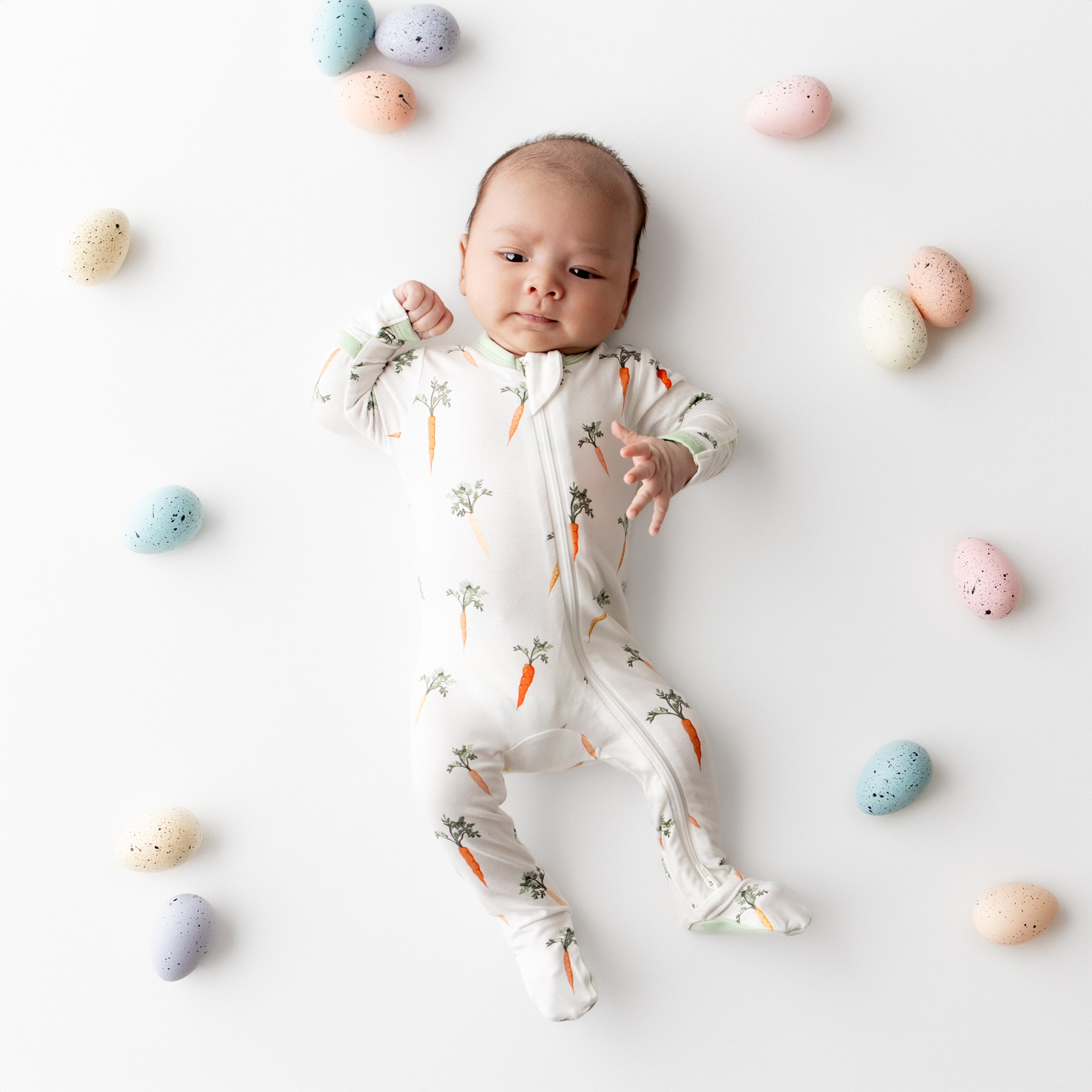 Newborn laying on a white surface wearing the Zippered footie in Carrot surrounded by colored easter eggs