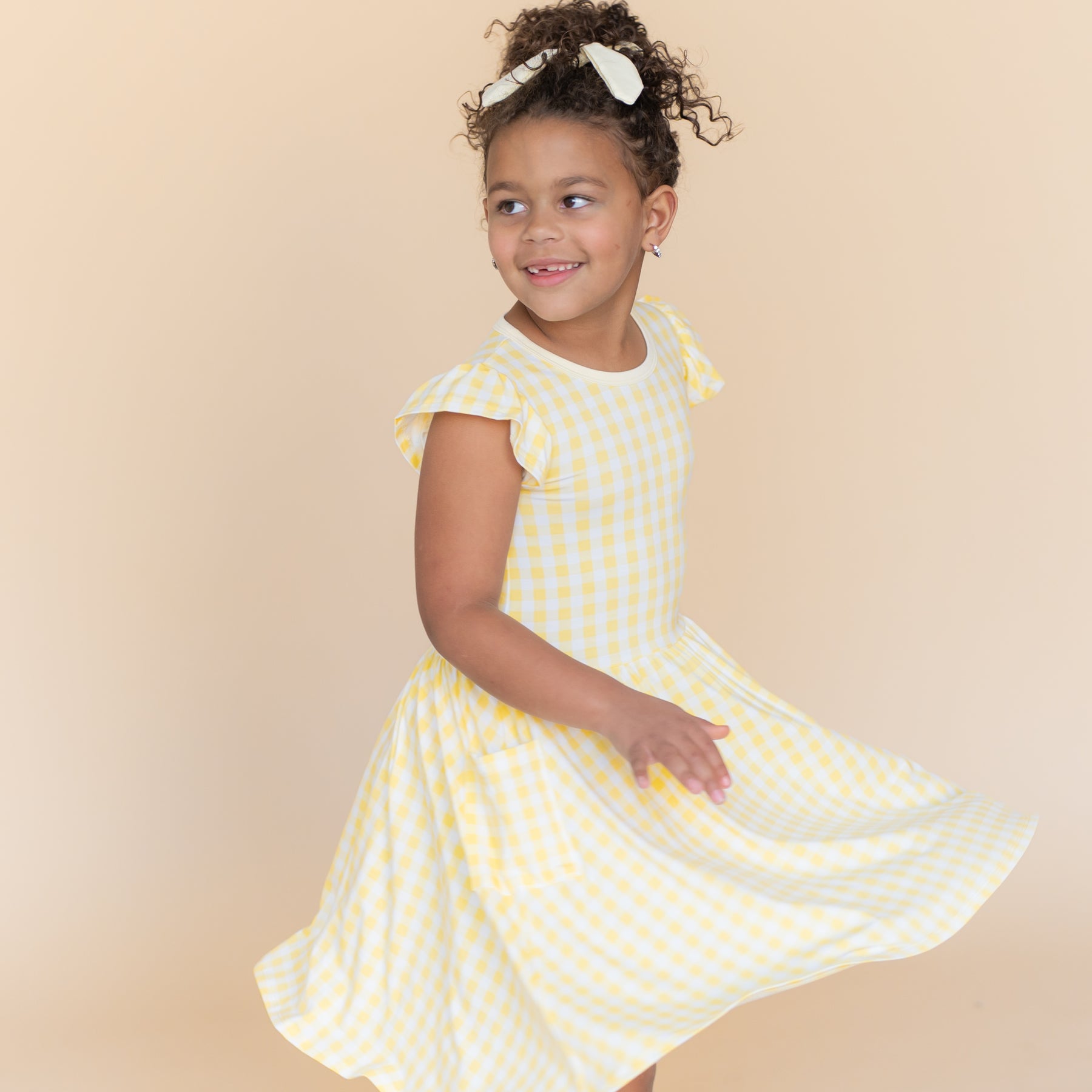 Young girl modeling the Pocket Dress in Gingham Chamomile twirling in front of a light taupe background