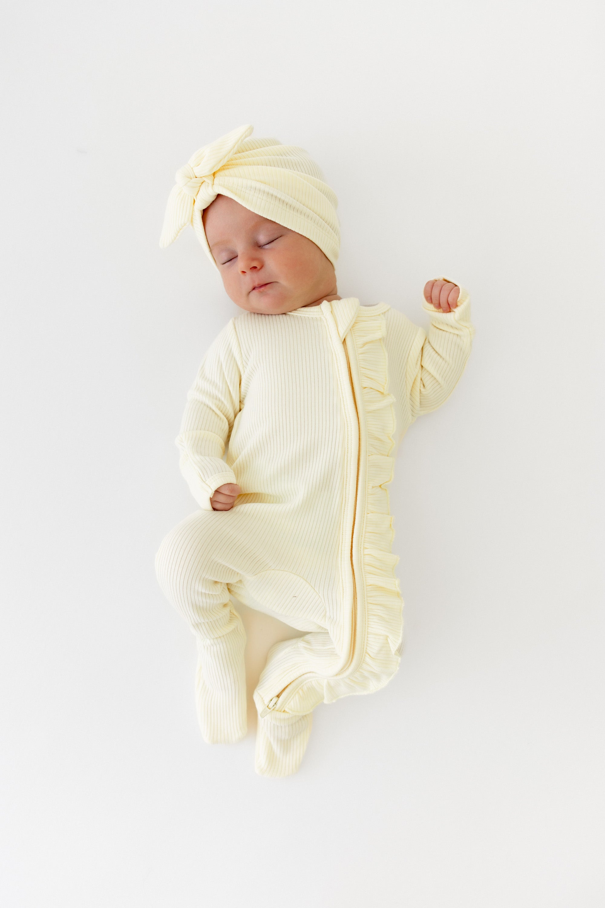 Sleeping infant wearing the Ribbed Ruffle Zipper Footie in Chamomile paired with a matching ribbed headwrap