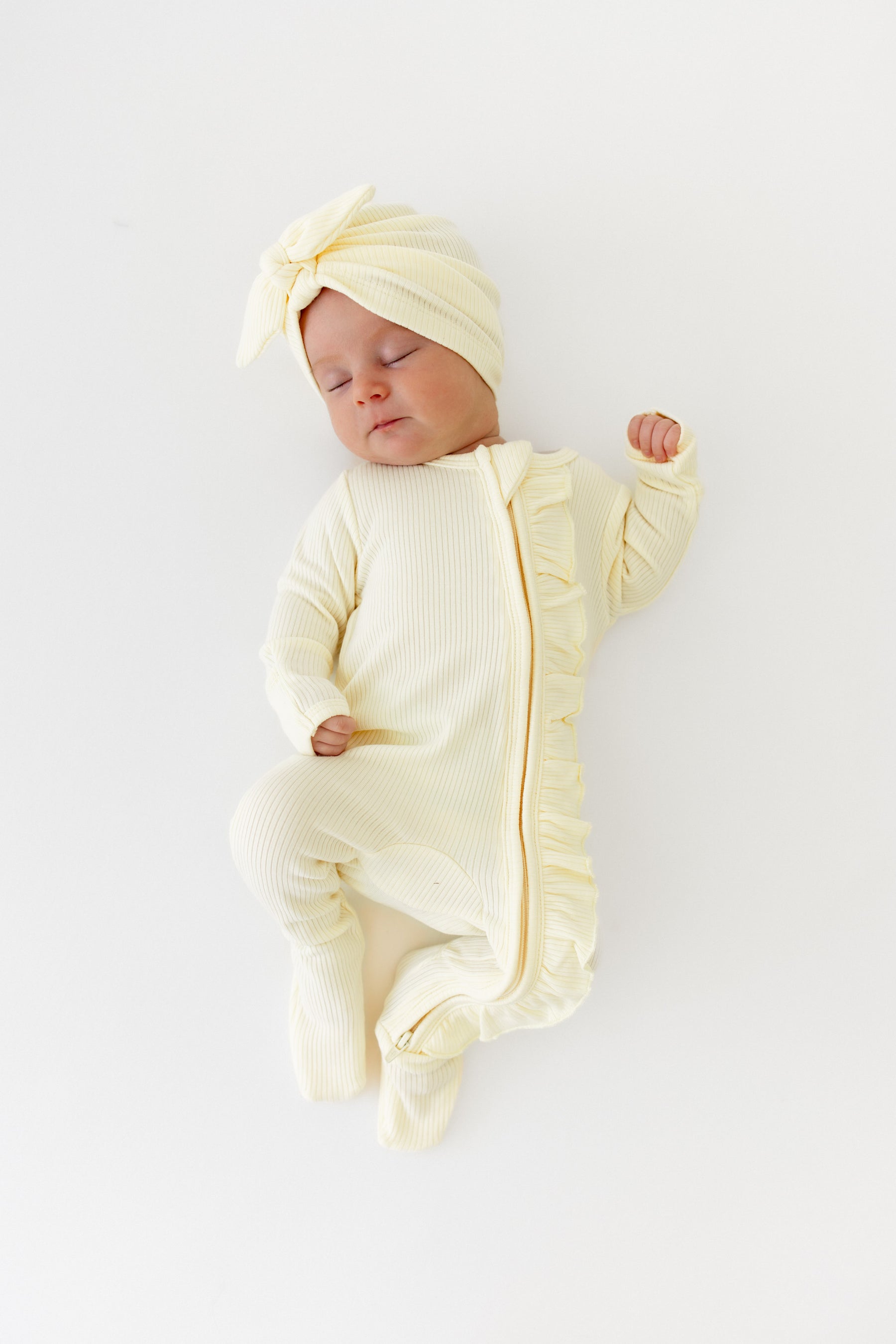 Sleeping infant wearing the Ribbed Ruffle Zipper Footie in Chamomile paired with a matching ribbed headwrap