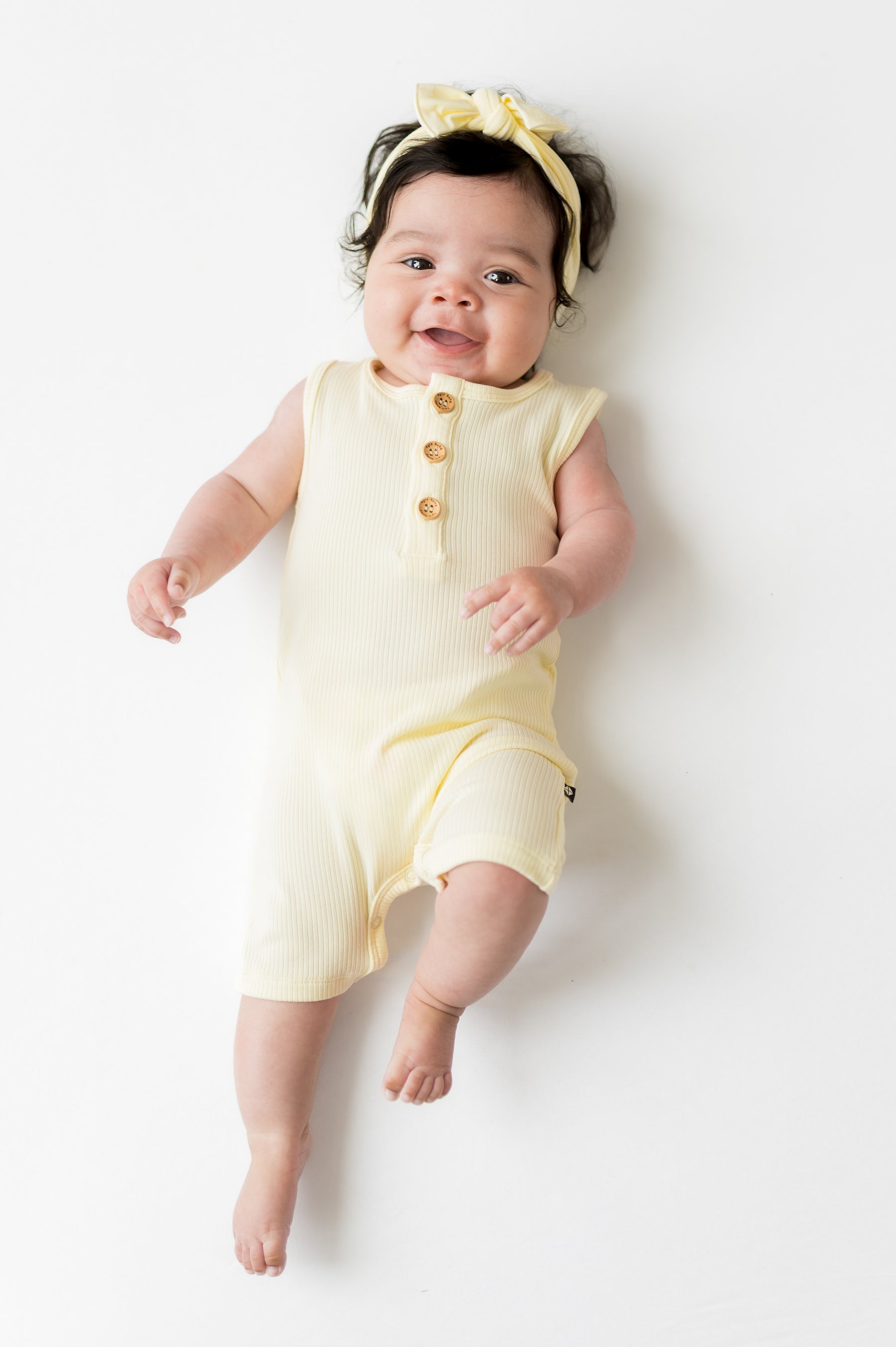 Smiling infant laying on a white surface wearing the Ribbed Sleeveless Shortall in Chamomile and matching bow
