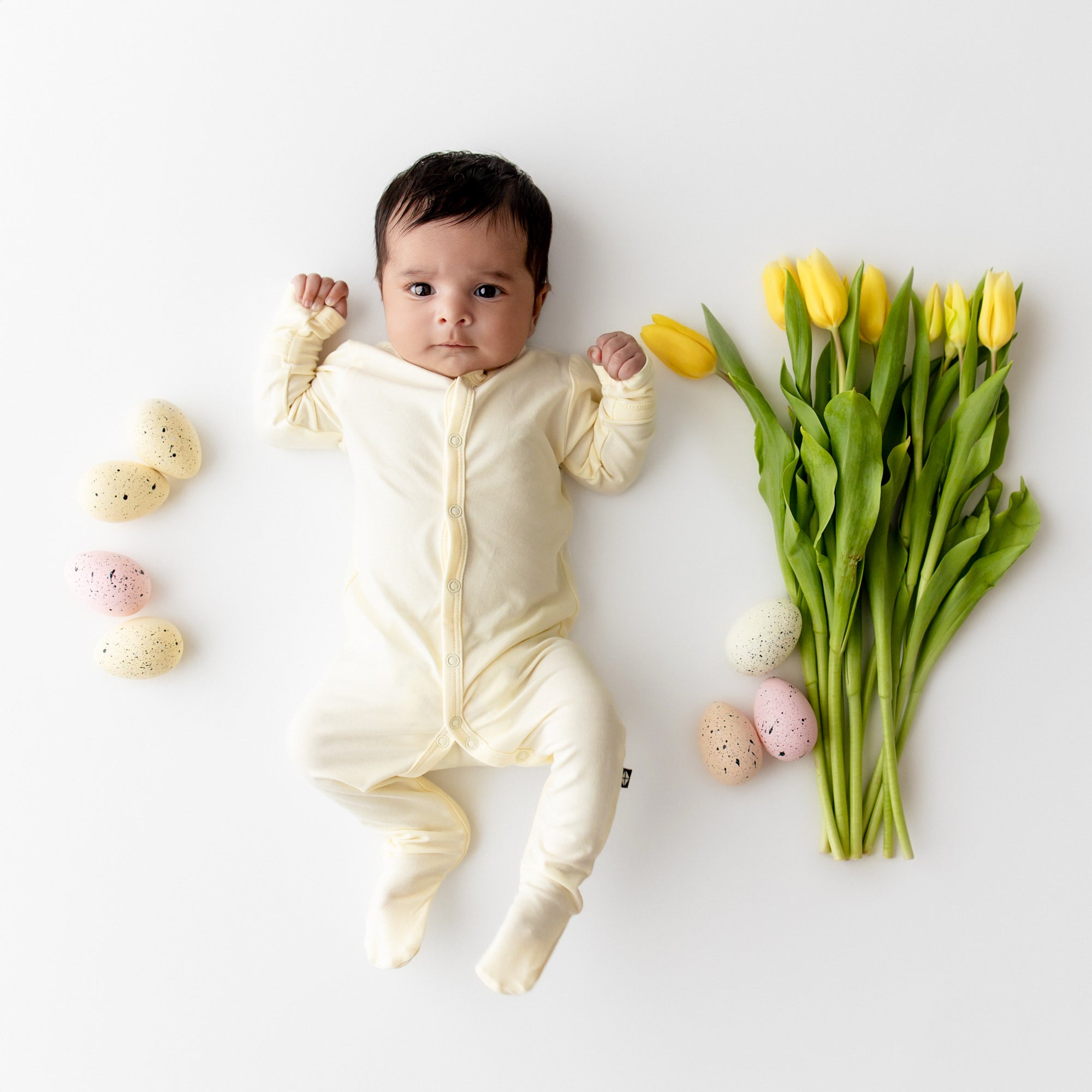 Baby wearing a pale yellow snap footie with easter egg and tulips around them
