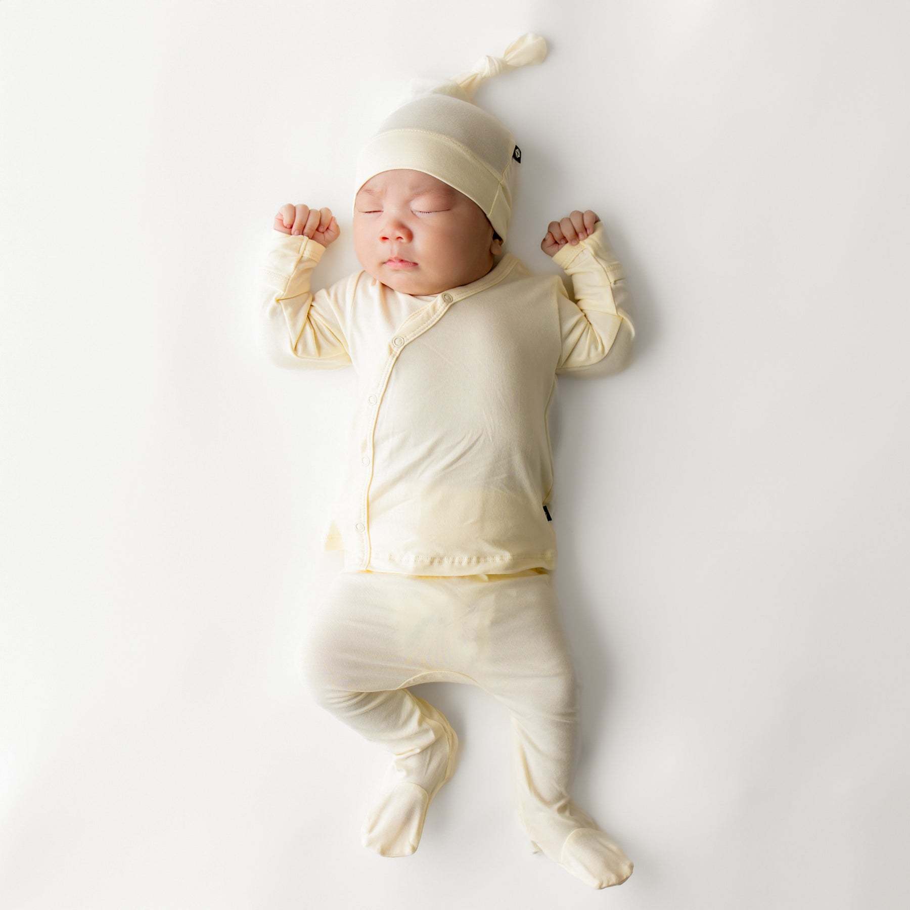 Sleeping newborn wearing the Take Me Home Set with Cap in Chamomile laying on a white surface