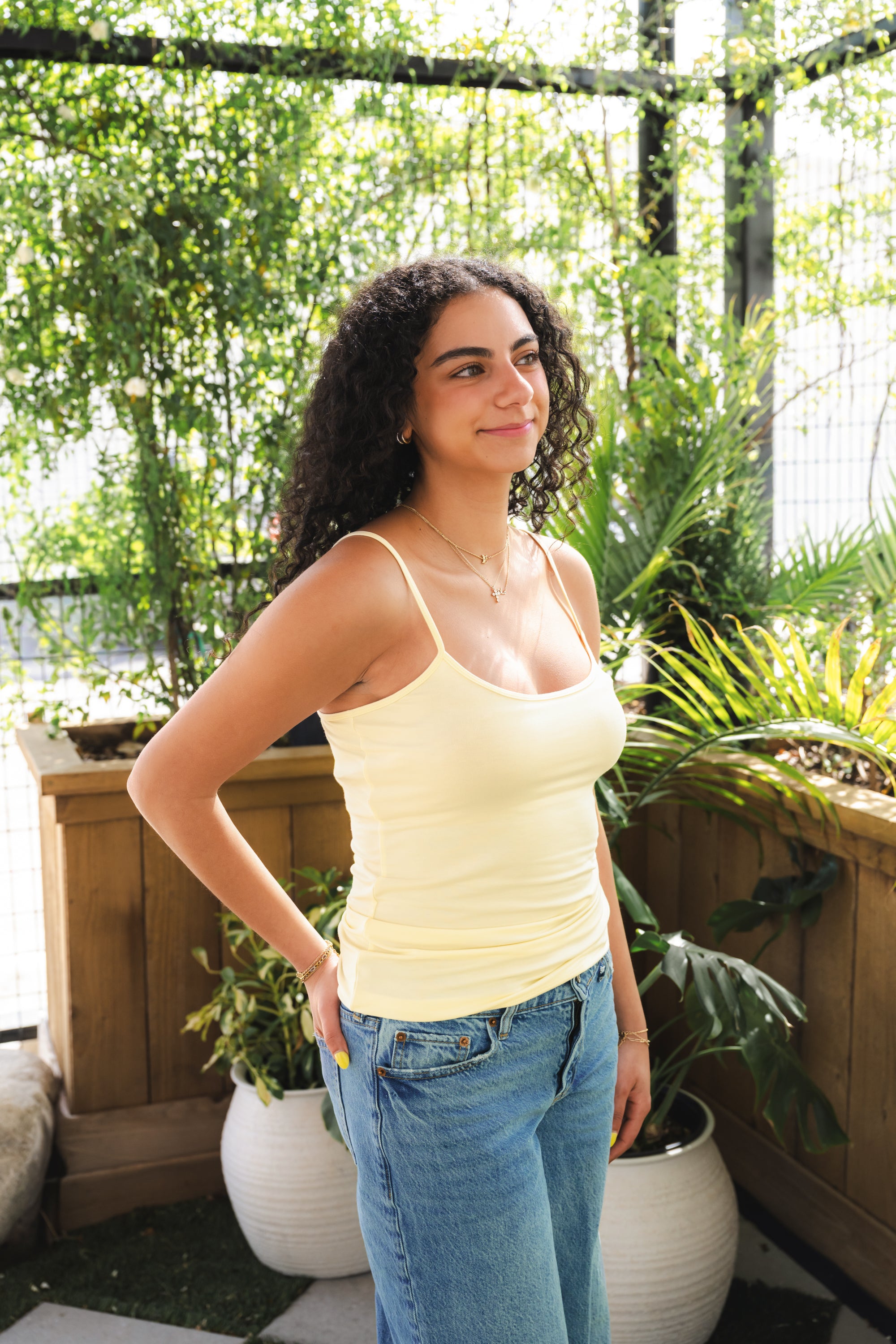 Woman wearing a yellow tank top and blue jeans standing in a garden setting.