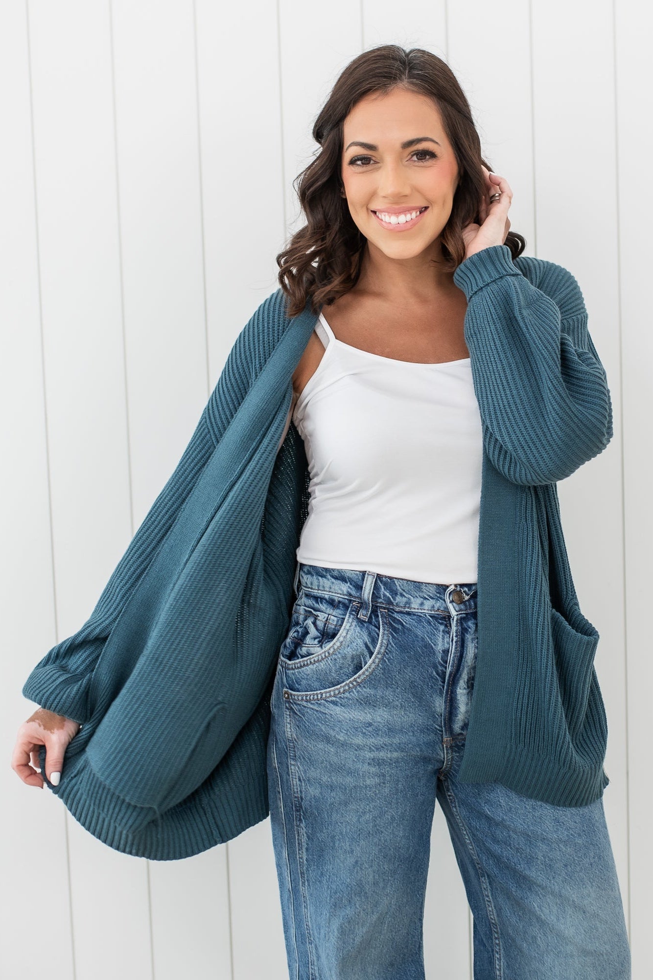 Smiling female model wearing the Chunky Knit Women's Oversized Cardigan in Atlantic paired with a white spaghetti strap top and medium wash jeans