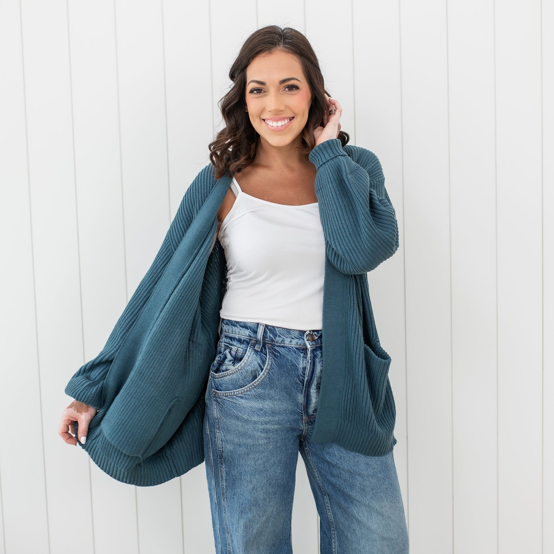 Smiling female model wearing the Chunky Knit Women's Oversized Cardigan in Atlantic paired with a white spaghetti strap top and medium wash jeans