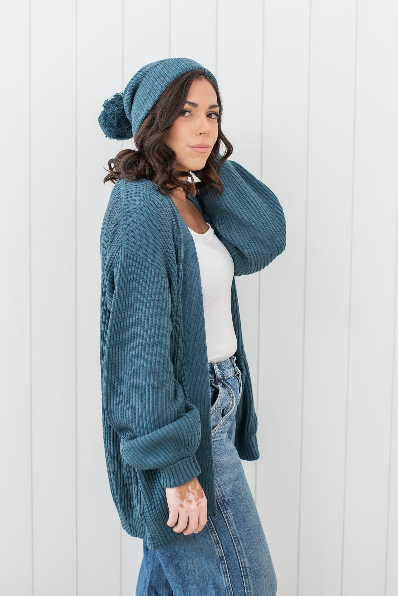 Female model standing in front of a white paneled wall wearing the Chunky Knit Women's Oversized Cardigan in Atlantic and matching chunky knit pom beanie