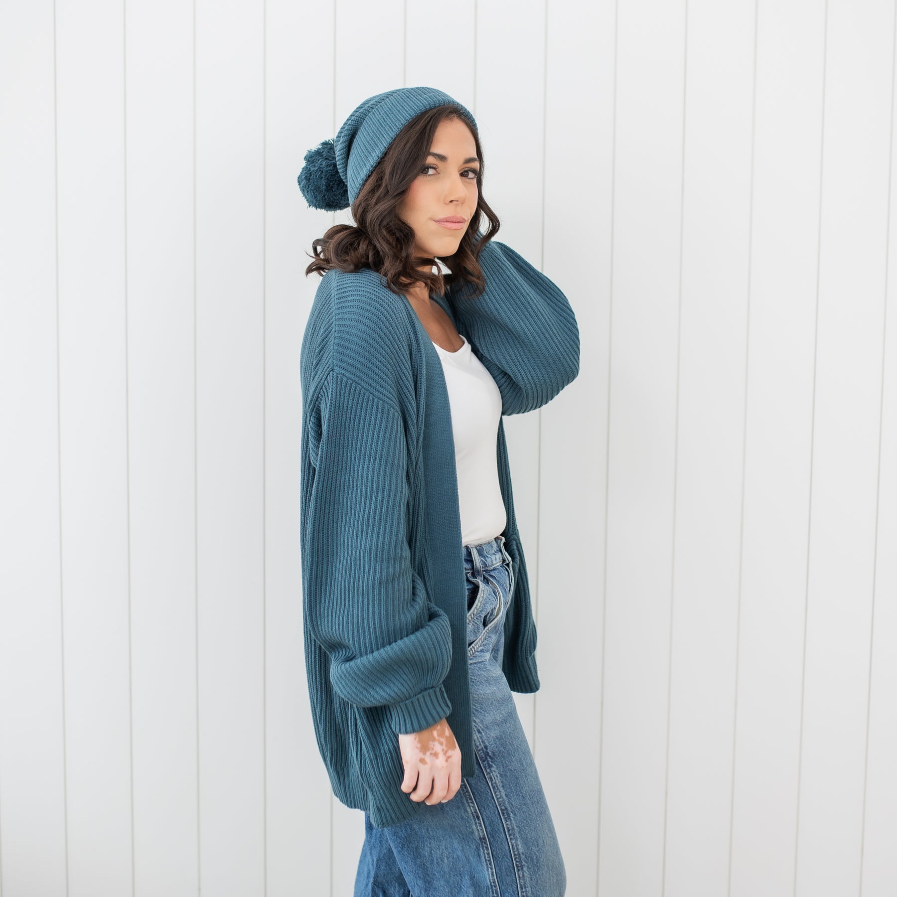 Female model standing in front of a white paneled wall wearing the Chunky Knit Women's Oversized Cardigan in Atlantic and matching chunky knit pom beanie