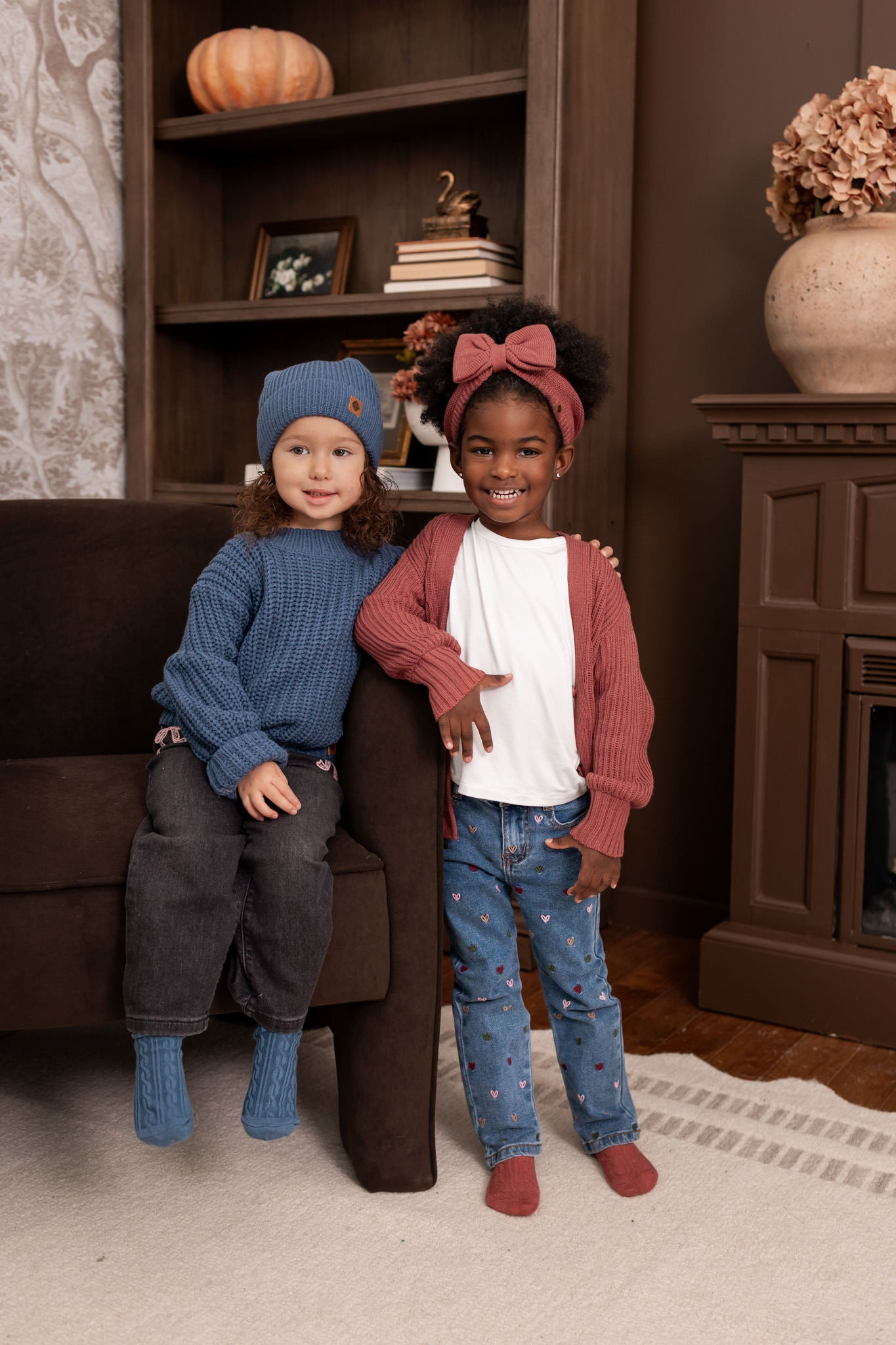 Two girls in a living room wearing Chunky Knit Sweater in Oxford with chunky knit pom beanie in oxford and chunky knit oversized cardigan in ember with chunky knit bow headband in ember
