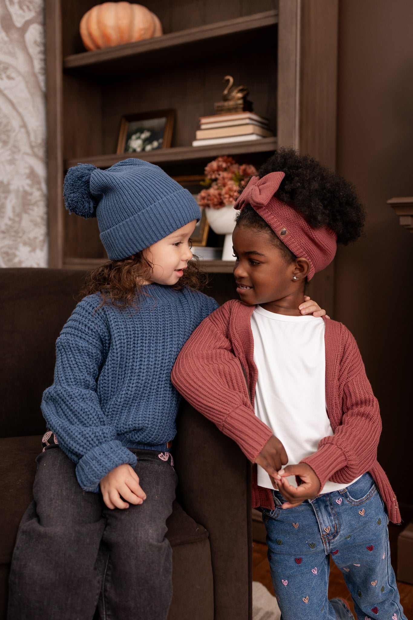 Two girls in talking to each other in a living room wearing Chunky Knit Sweater in Oxford with chunky knit pom beanie in oxford and chunky knit oversized cardigan in ember with chunky knit bow headband in ember