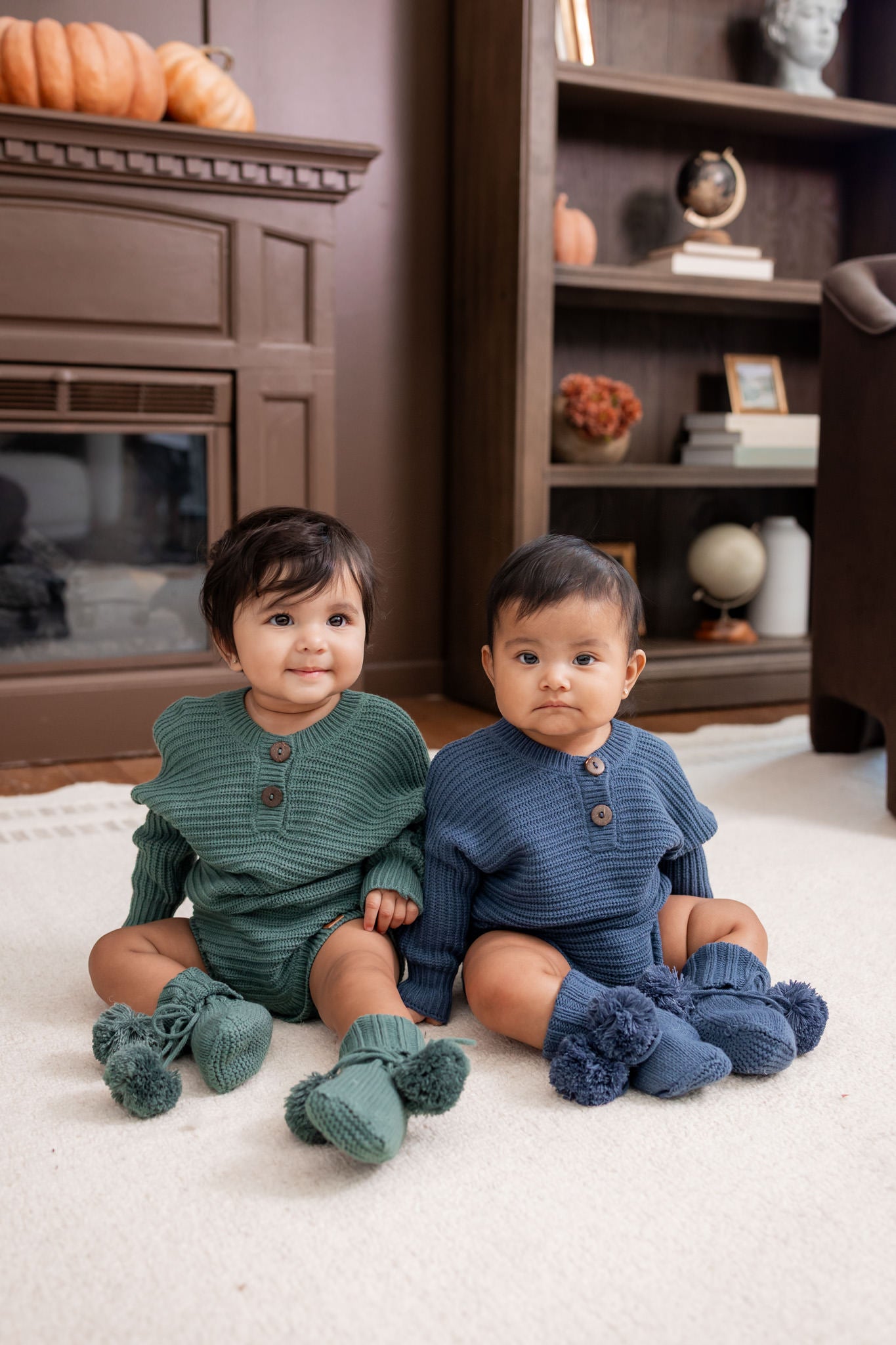 Two babies sitting on white carpet in a living room wearing Chunky Knit Romper in Pine and oxford and knit booties in pine and oxford