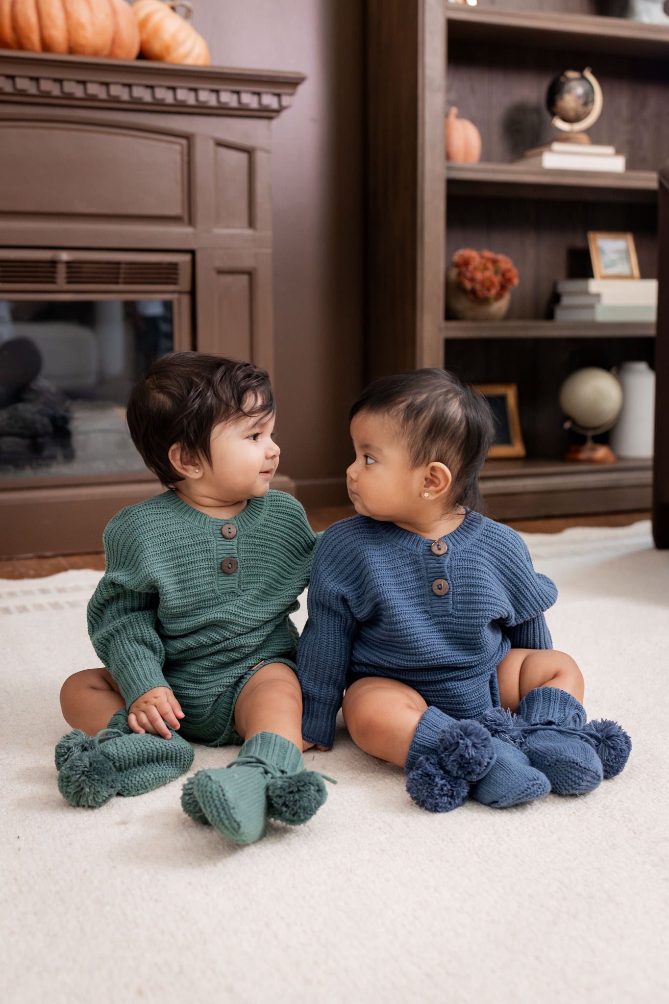 Two babies looking at each other sitting on white carpet in a living room wearing Chunky Knit Romper in Pine and oxford and knit booties in pine and oxford