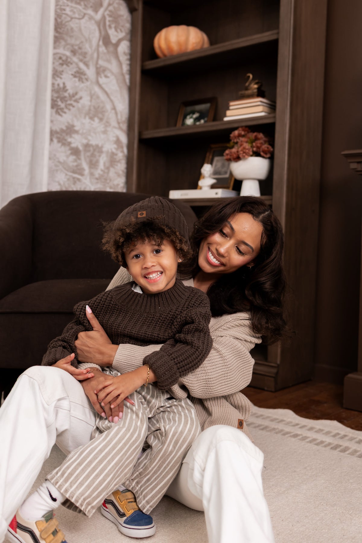 Modal wearing Chunky Knit Women's Cropped Cardigan in Almond with child wearing chunky knit ribbed beanie and Chunky knit sweater in espresso sitting in al living room