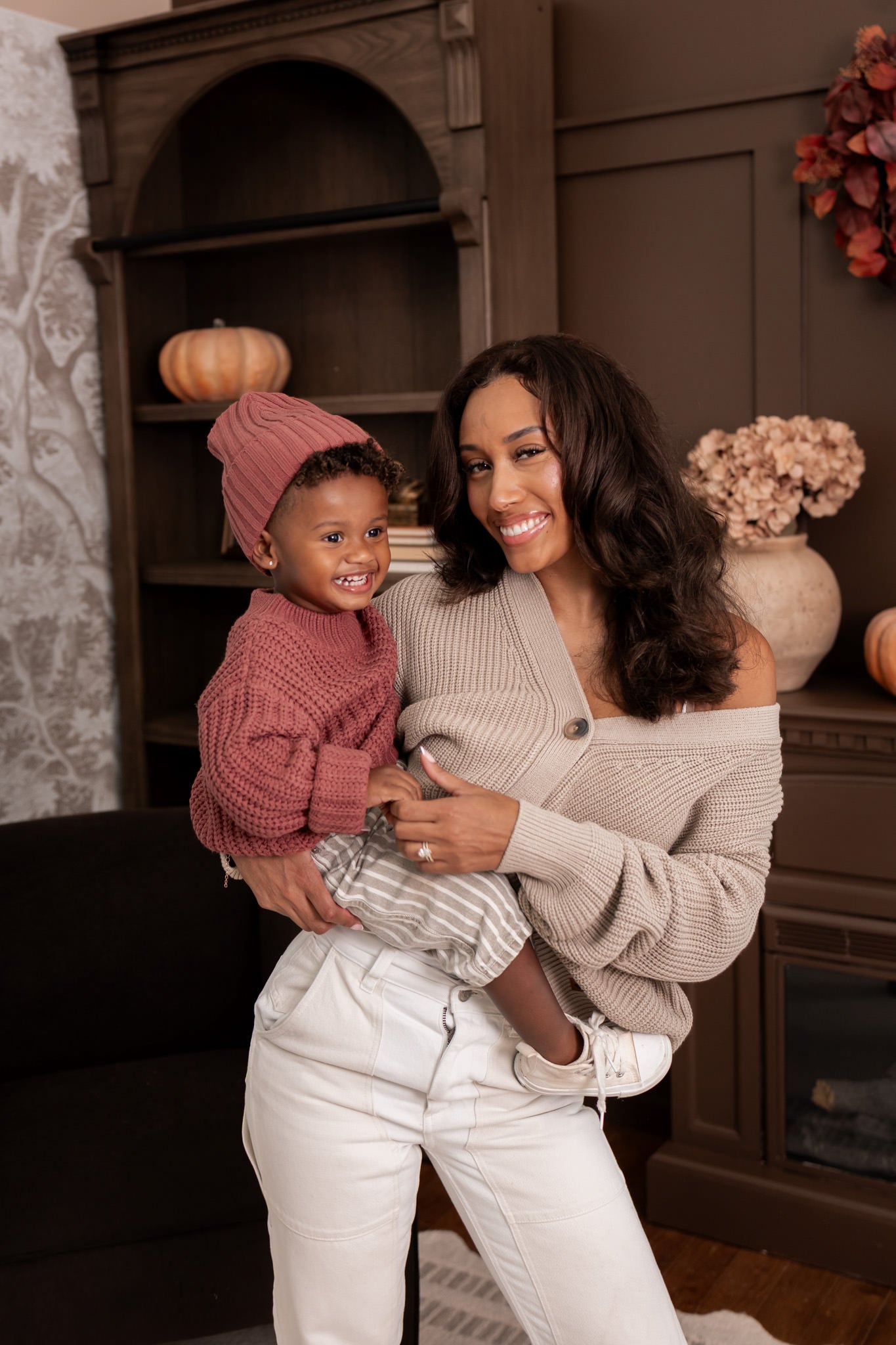Modal wearing Chunky Knit Women's Cropped Cardigan in Almond with child wearing chunky knit ribbed beanie and Chunky knit sweater in espresso sitting in al living room