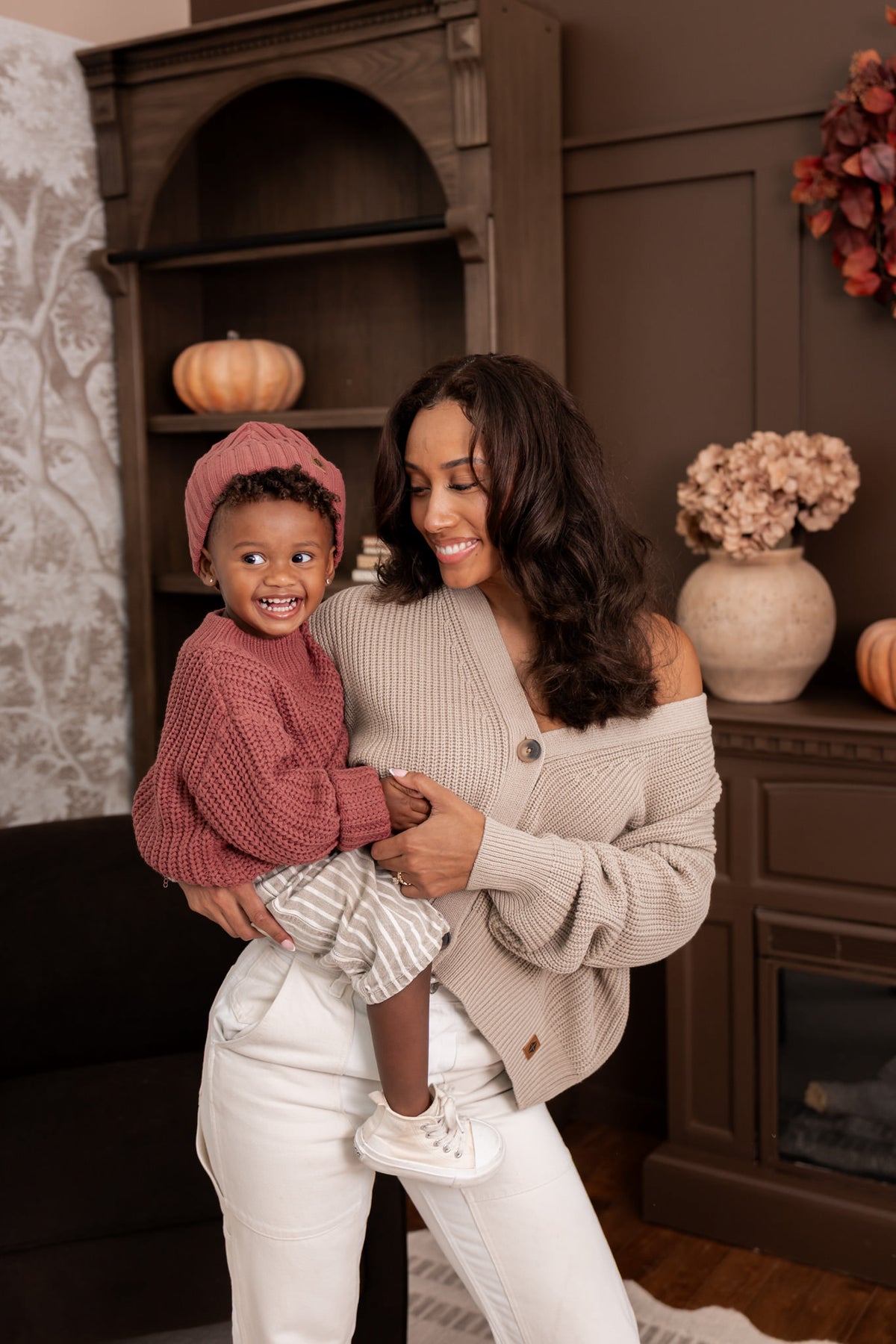Modal wearing Chunky Knit Women's Cropped Cardigan in Almond with child wearing chunky knit ribbed beanie and Chunky knit sweater in espresso sitting in al living room