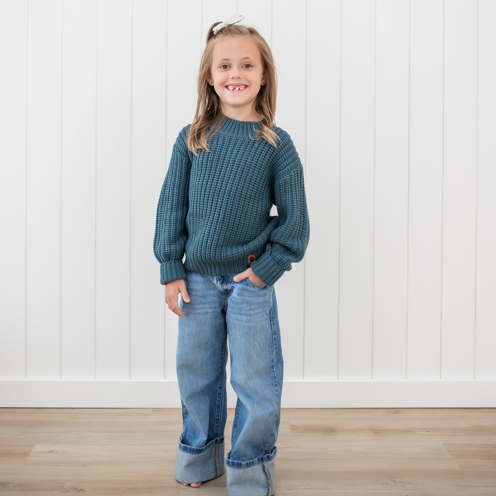 Smiling girl wearing the Chunky Knit Sweater in Atlantic paired with medium wash jeans standing in front of a white paneled wall