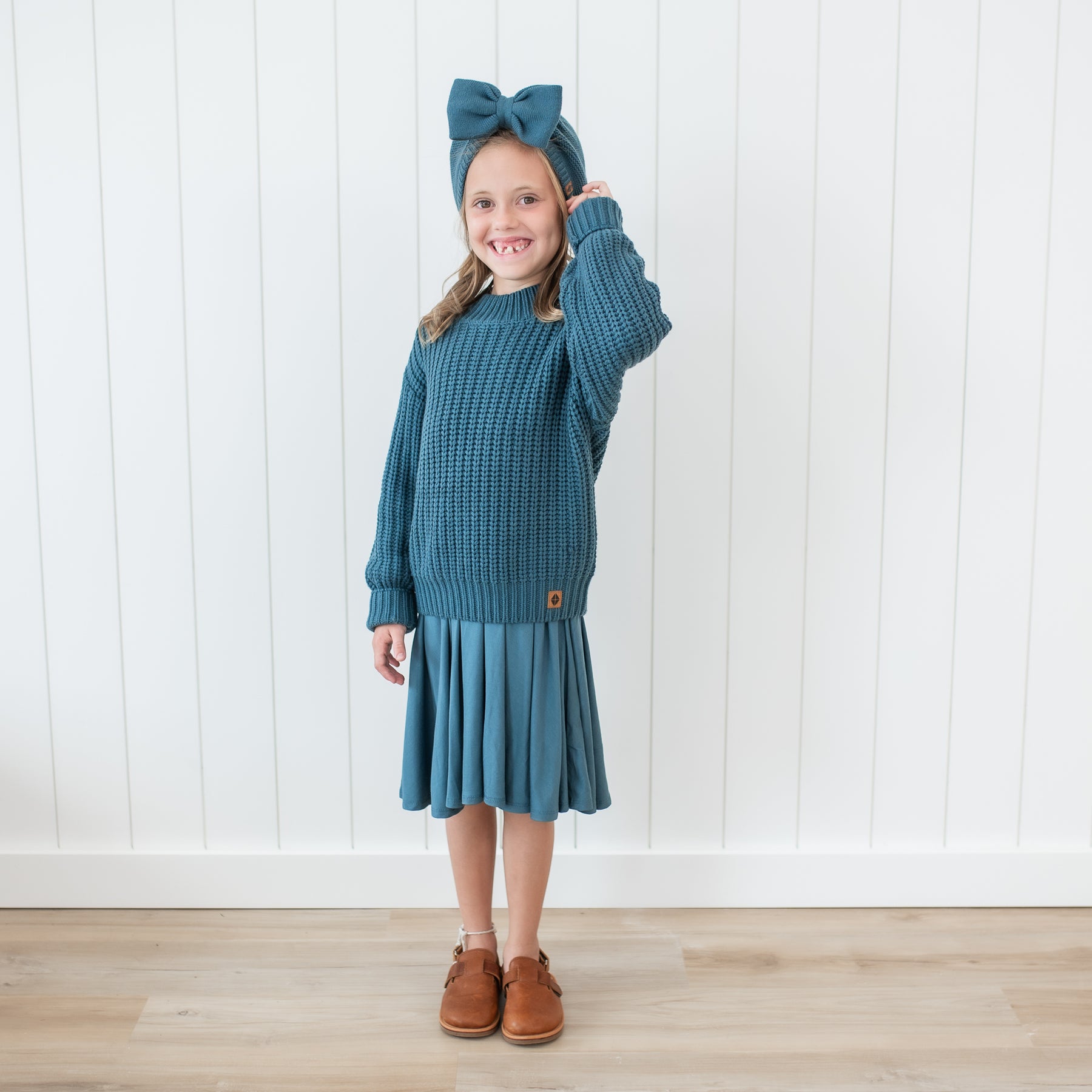 Young girl standing in front of a white paneled wall wearing the Chunky Knit Bow Headband in Atlantic and matching Twirl Dress and Chunky Knit Sweater