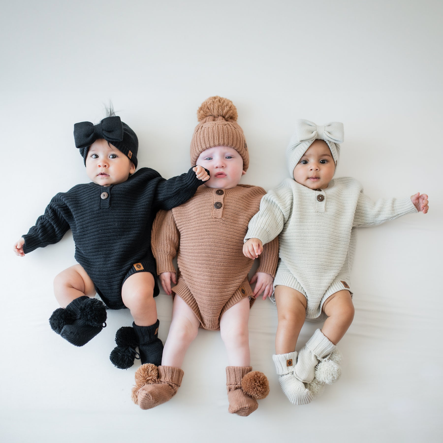 Three infants laying side by side wearing the Chunky Knit Romper in Latte, Oat and Midnight with matching knit booties