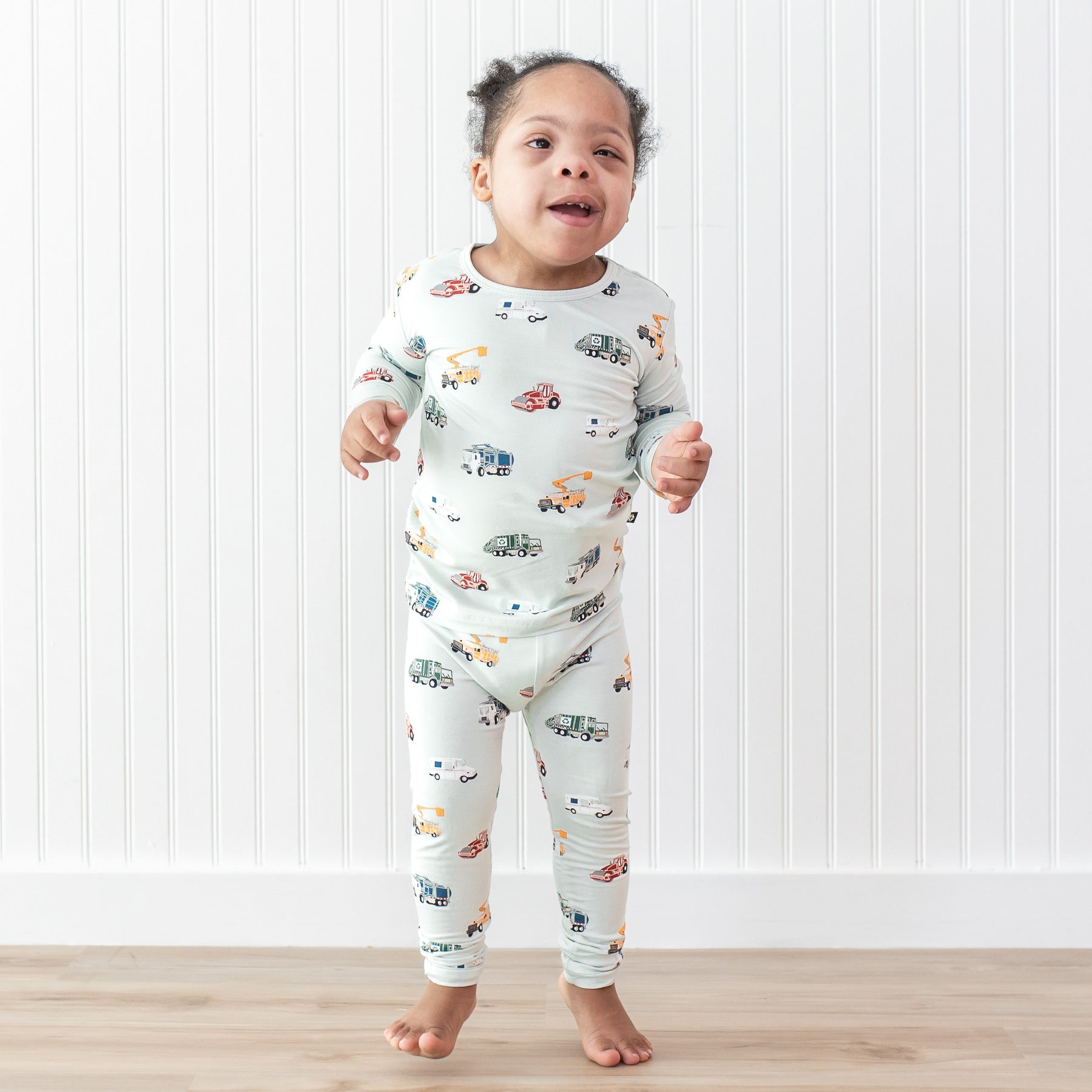 Young child smiling wearing the Long Sleeve Pajamas in City Vehicles standing in front of a white paneled wall