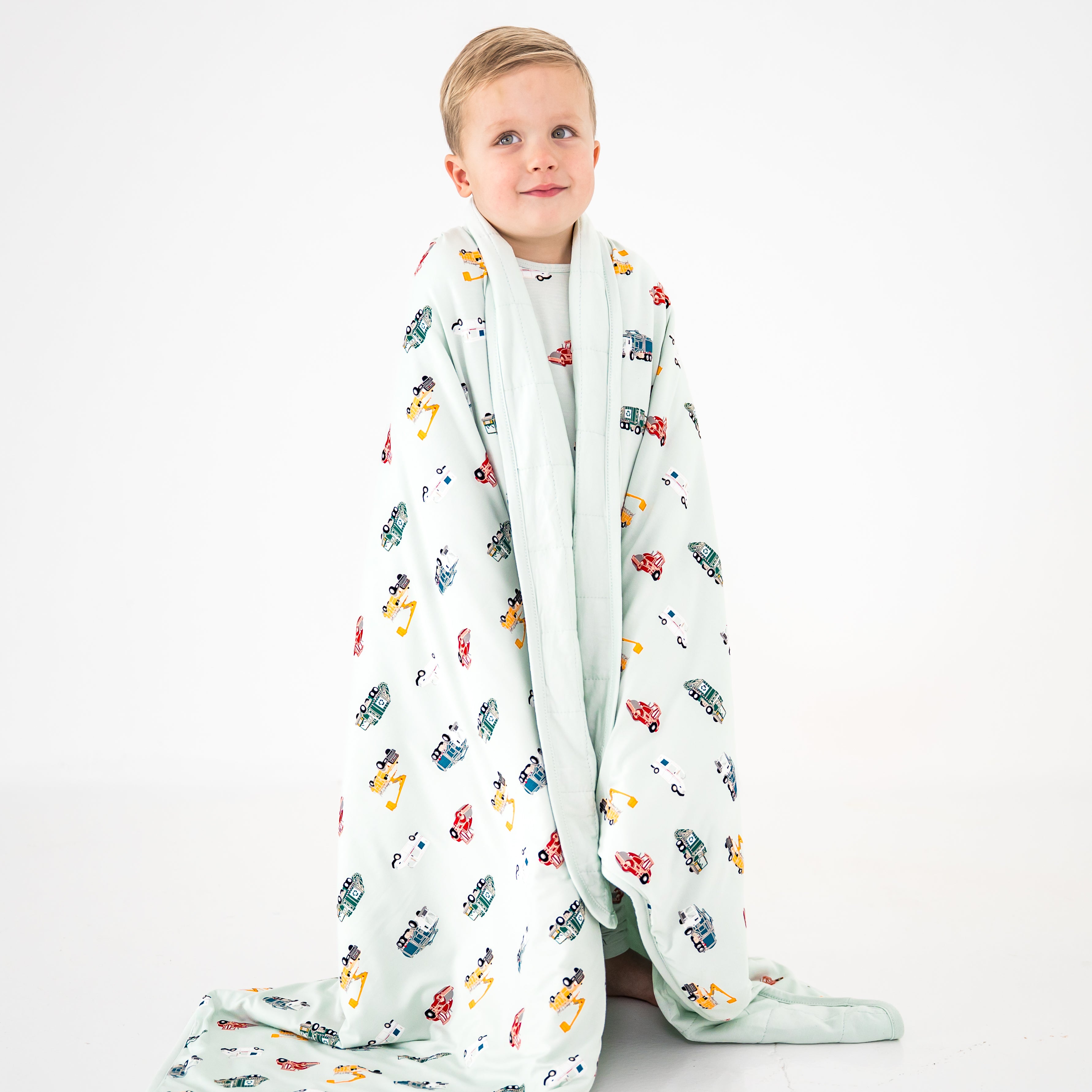Young boy standing in front of a white background with the soft and breathable Toddler Blanket in City Vehicles 1.0 around his shoulders