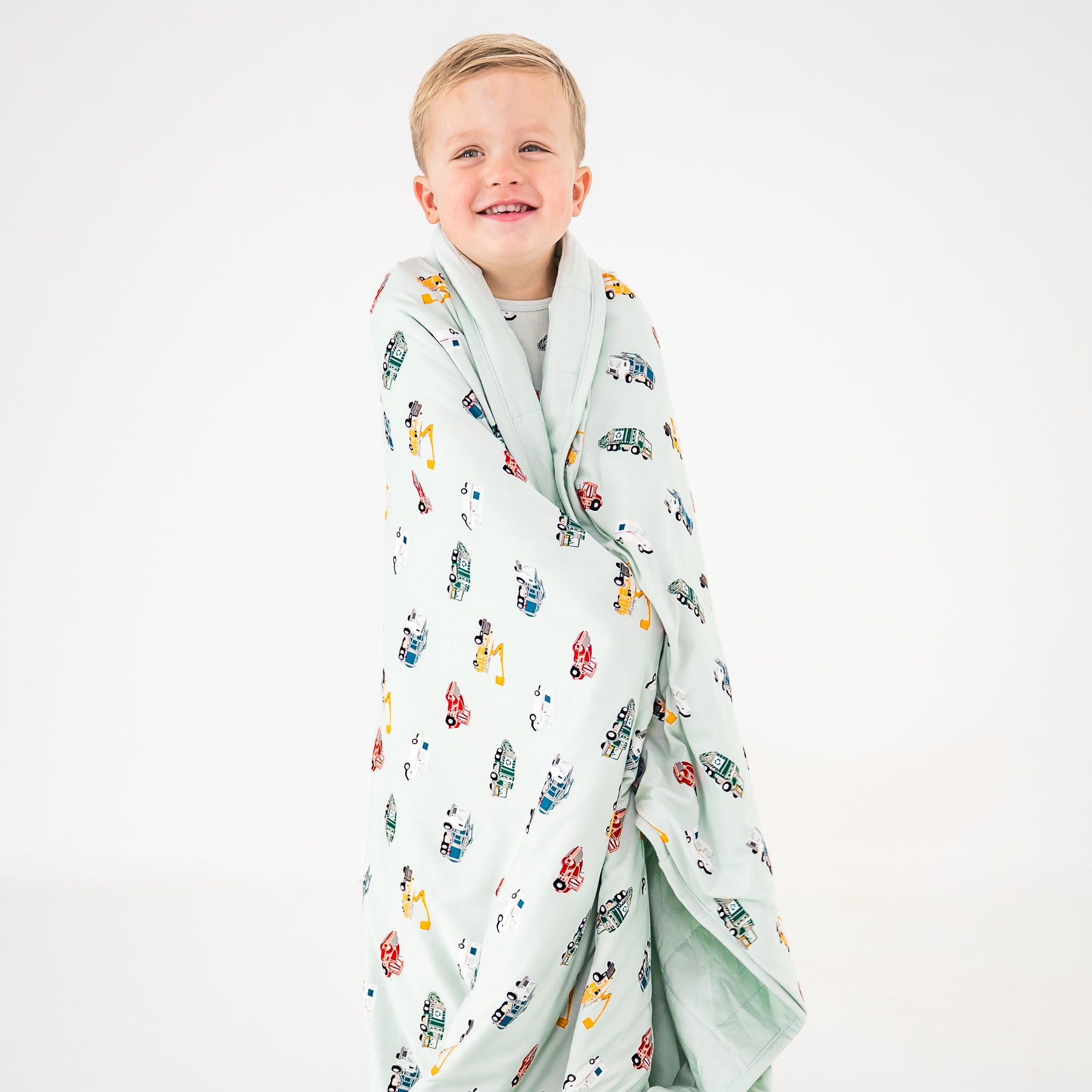 Young smiling boy standing in front of a white background with the Toddler Blanket in City Vehicles 1.0 around his shoulders