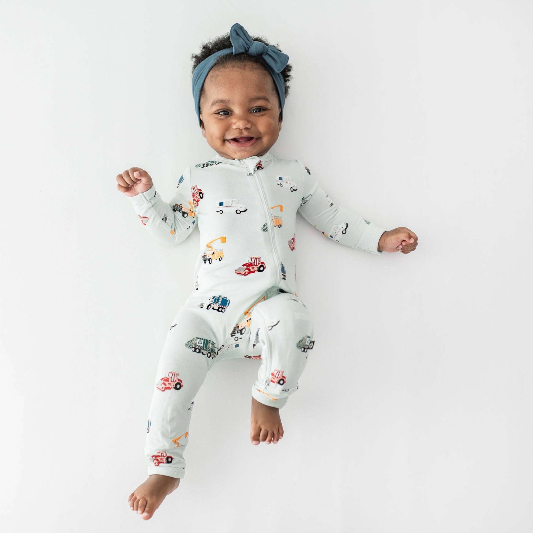 Young toddler laying on a white surface wearing the Zippered Romper in City Vehicles paired with an Atlantic Knotted Bow Headband