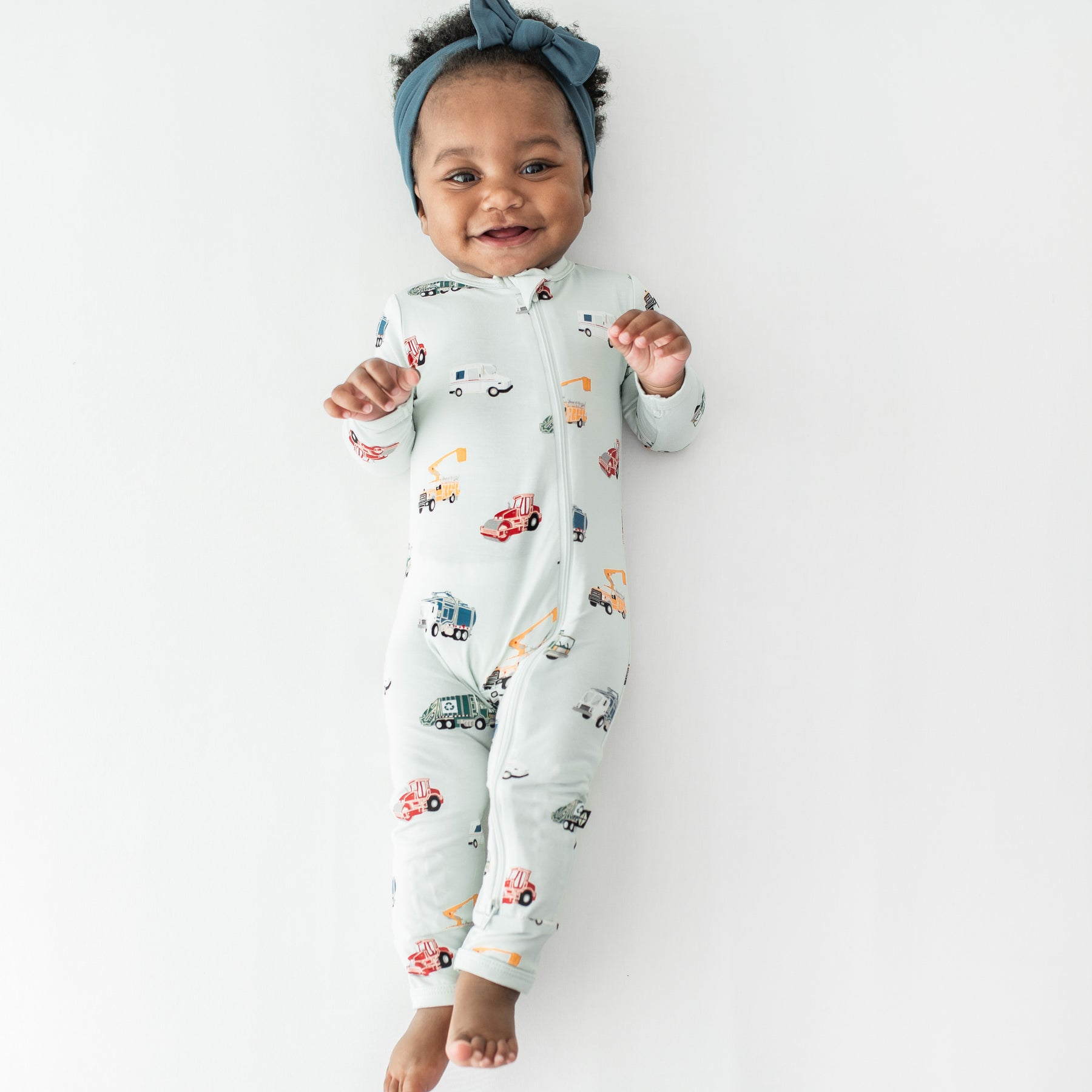 Young toddler girl laying stretched on a white surface wearing the Zippered Romper in City Vehicles and muted blue knotted bow headband