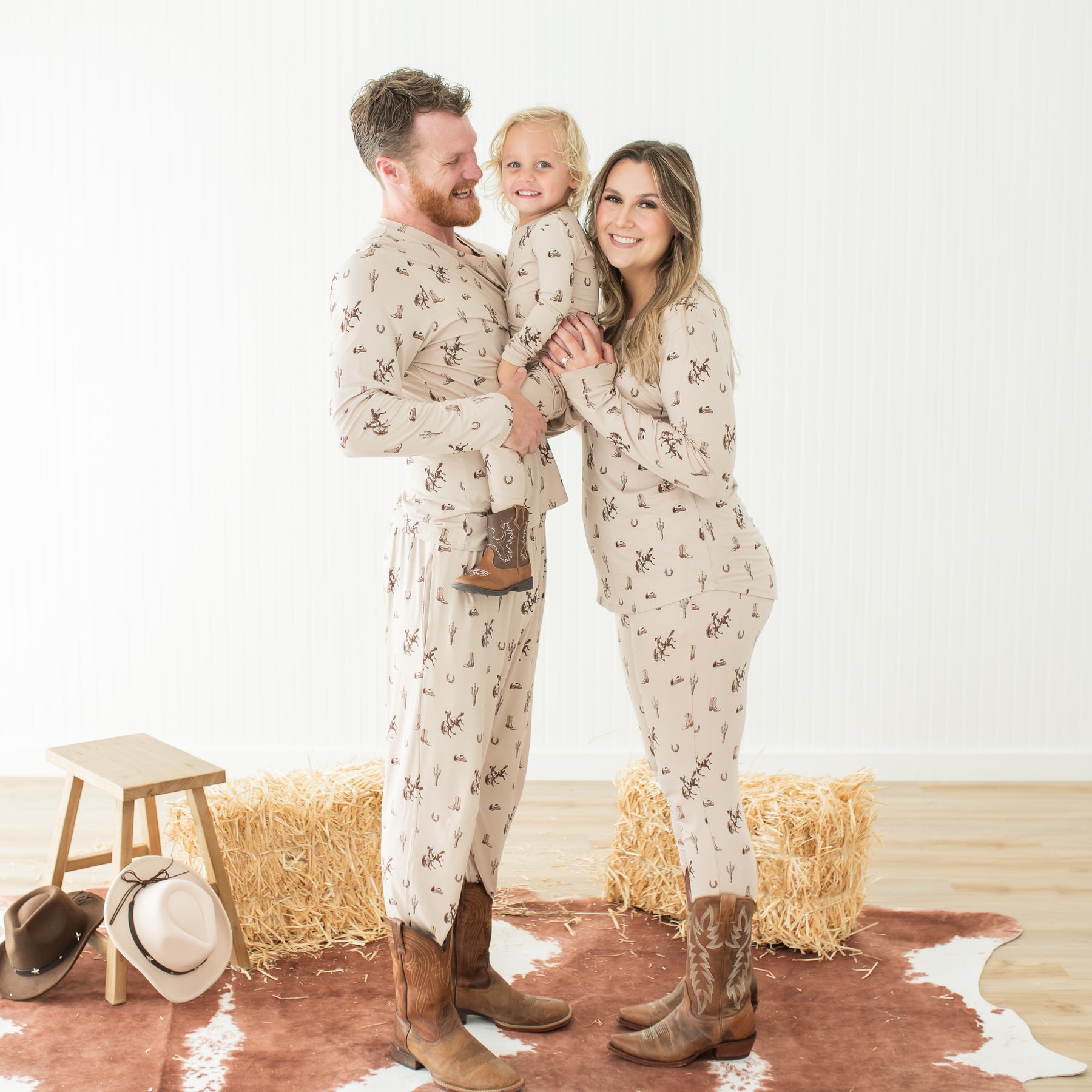 Family of three matching in Classic Cowboy pajamas and cowboy boots standing in front of hay bales 