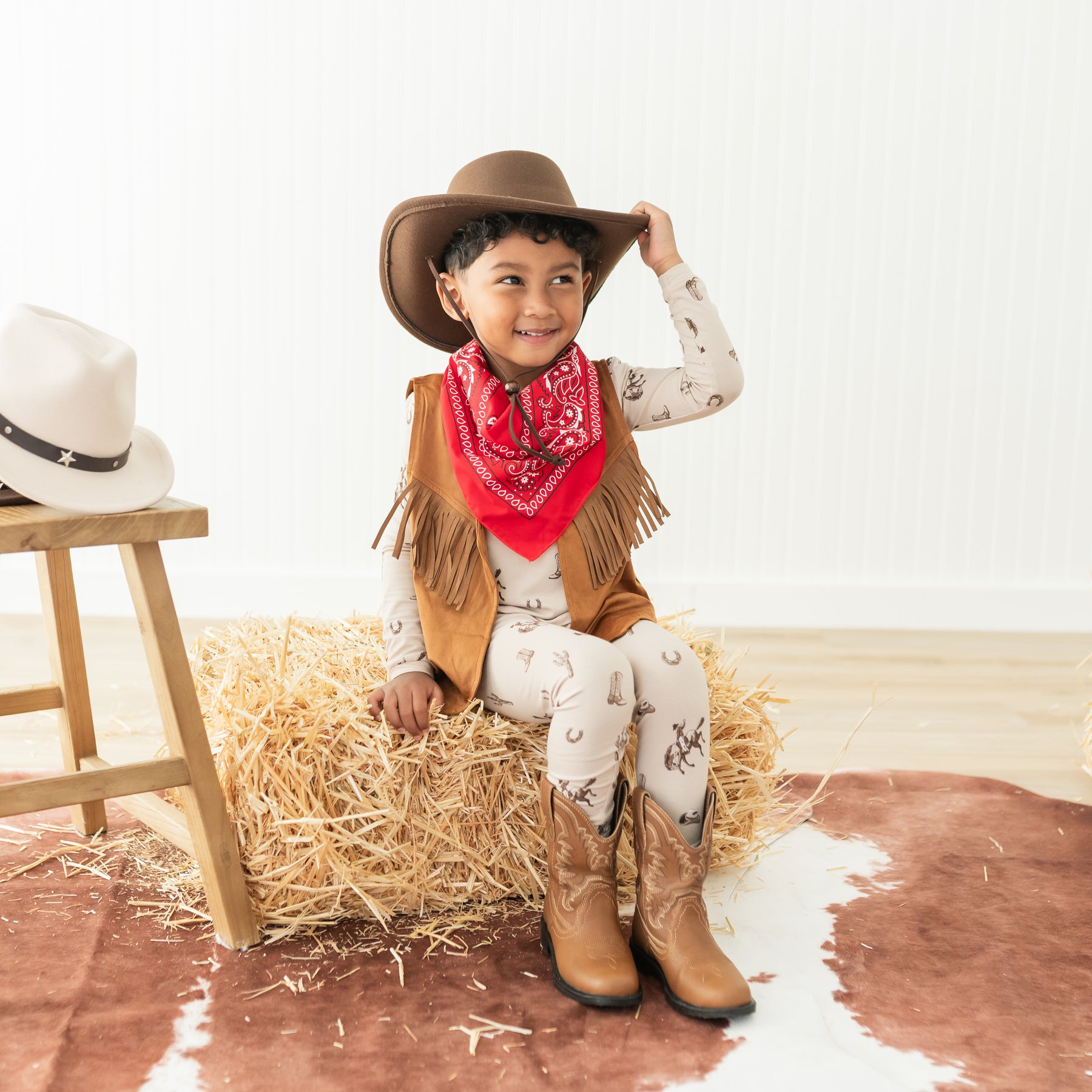 Young boy sitting on a hay bale wearing the Long Sleeve Pajamas in Classic Cowboy with a brown western vest, a cowboy hat and cowboy boots
