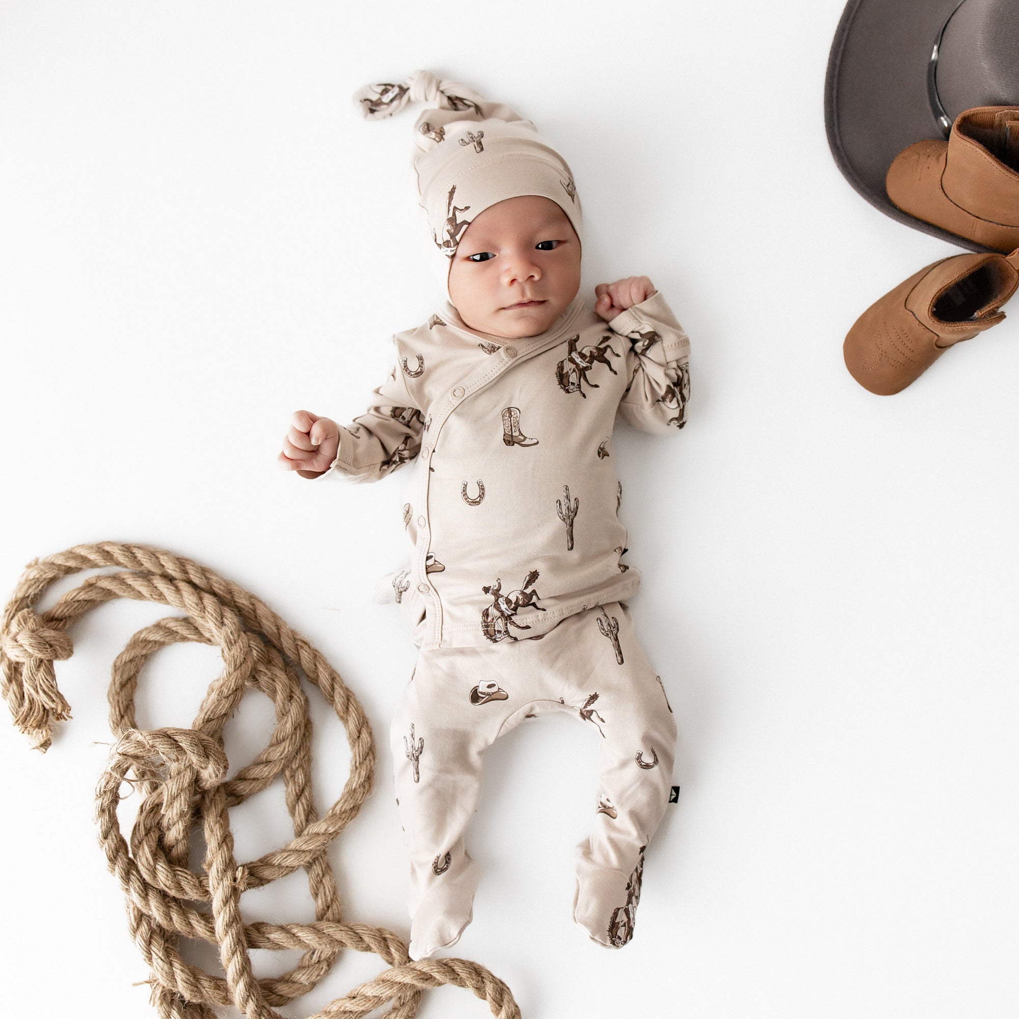 Newborn laying on a cream surface wearing the soft Take Me Home Set with Cap in Classic Cowboy from Kyte Baby with a robe, brown cowboy boot and cowboy hat around him