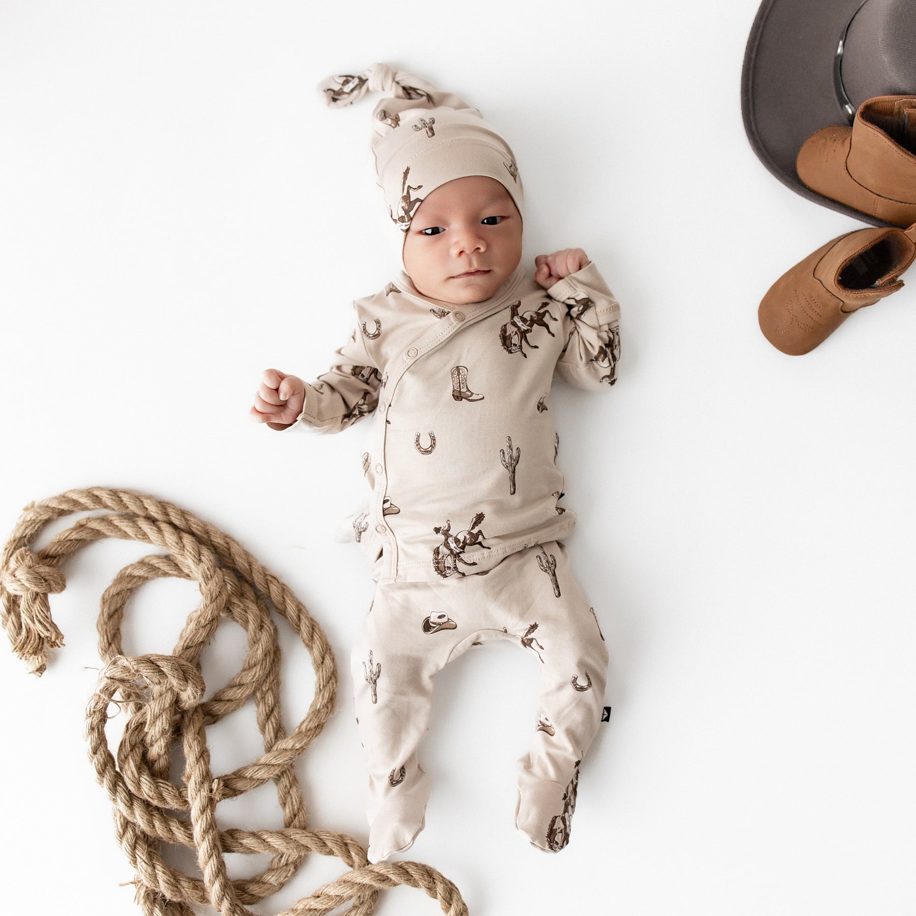 Newborn laying on a cream surface wearing the soft Take Me Home Set with Cap in Classic Cowboy from Kyte Baby with a robe, brown cowboy boot and cowboy hat around him