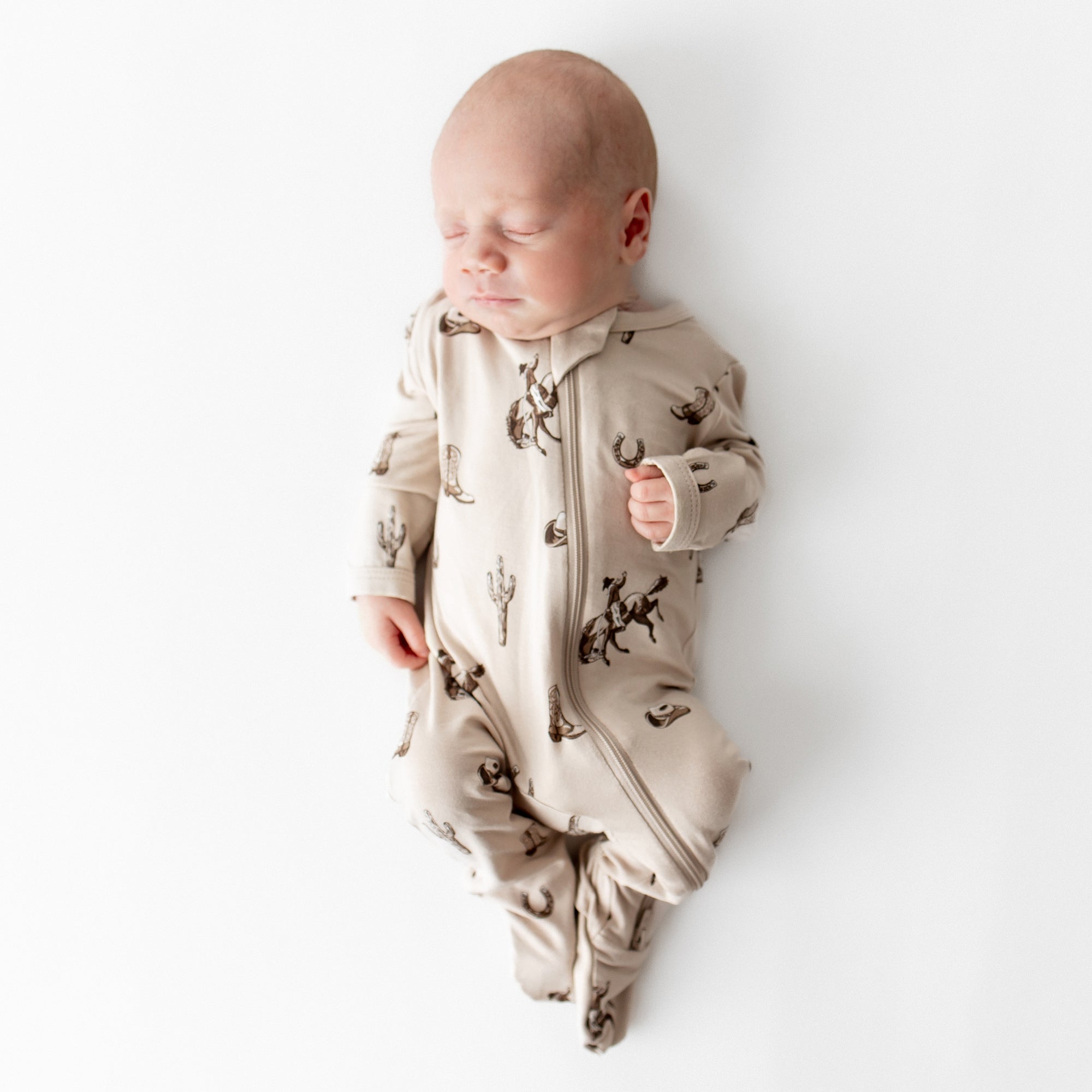 Sleeping newborn wearing the soft and breathable Zippered Footie in Classic Cowboy from Kyte Baby laying on a light cream surface