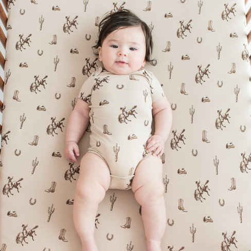 Infant wearing the Bodysuit in Classic Cowboy laying in a crib on a matching crib sheet