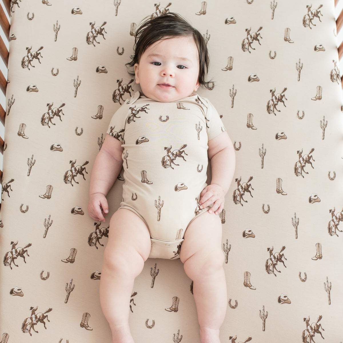 Infant wearing the Bodysuit in Classic Cowboy laying in a crib on a matching crib sheet