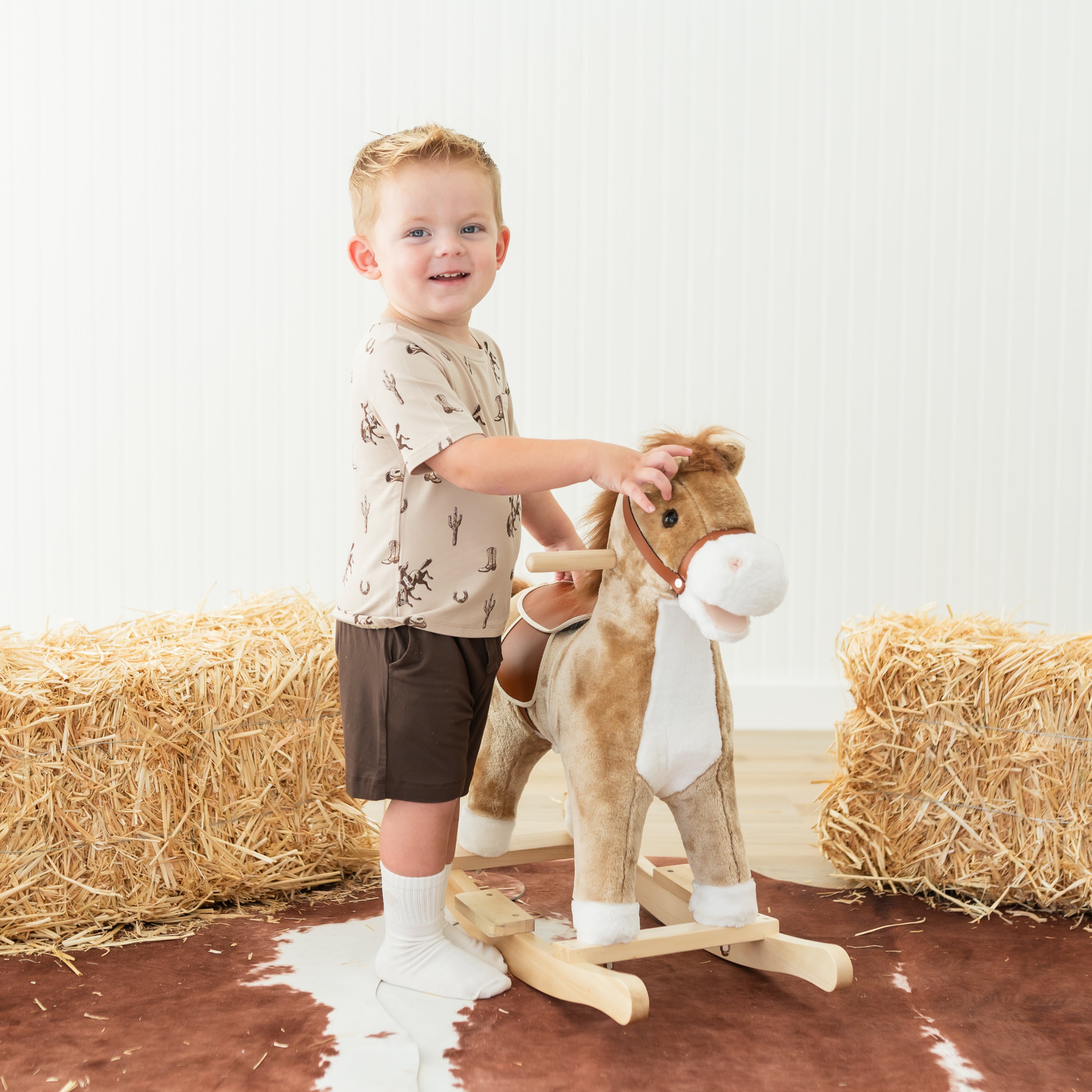 Young boy standing beside a toy horse rocker wearing the Toddler Crew Neck Tee in Classic Cowboy and brown shorts