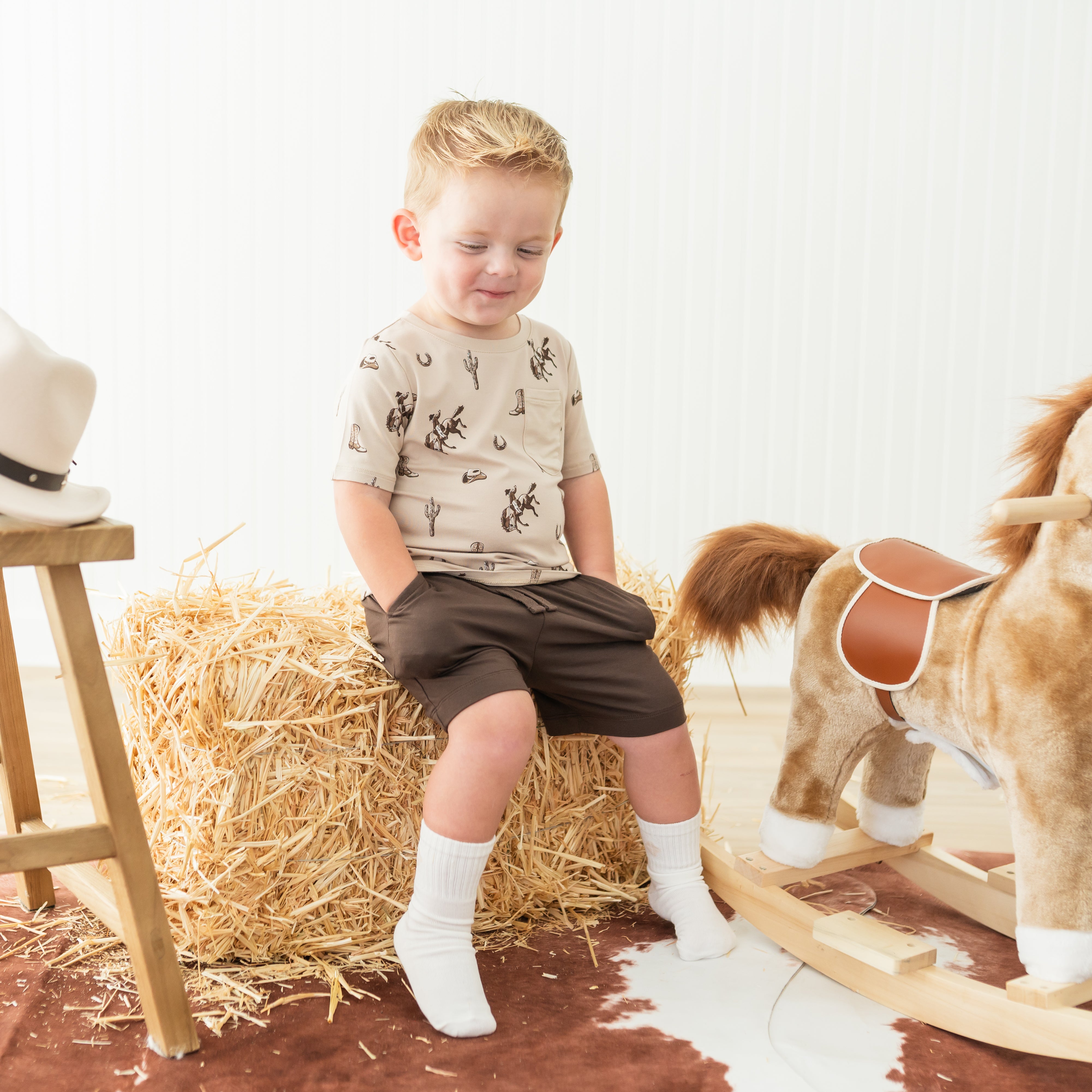 Smiling boy sitting on a hay bale wearing the Toddler Crew Neck Tee in Classic Cowboy paired with brown drawstring shorts