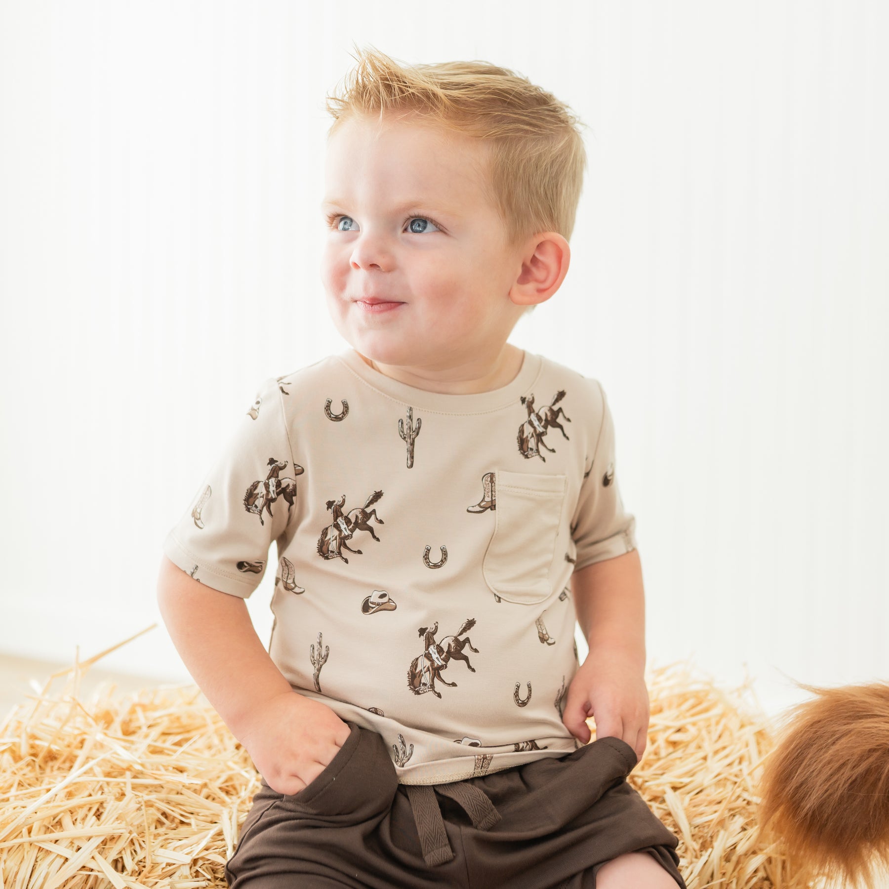 Smiling boy sitting on a hay bale wearing the Toddler Crew Neck Tee in Classic Cowboy 