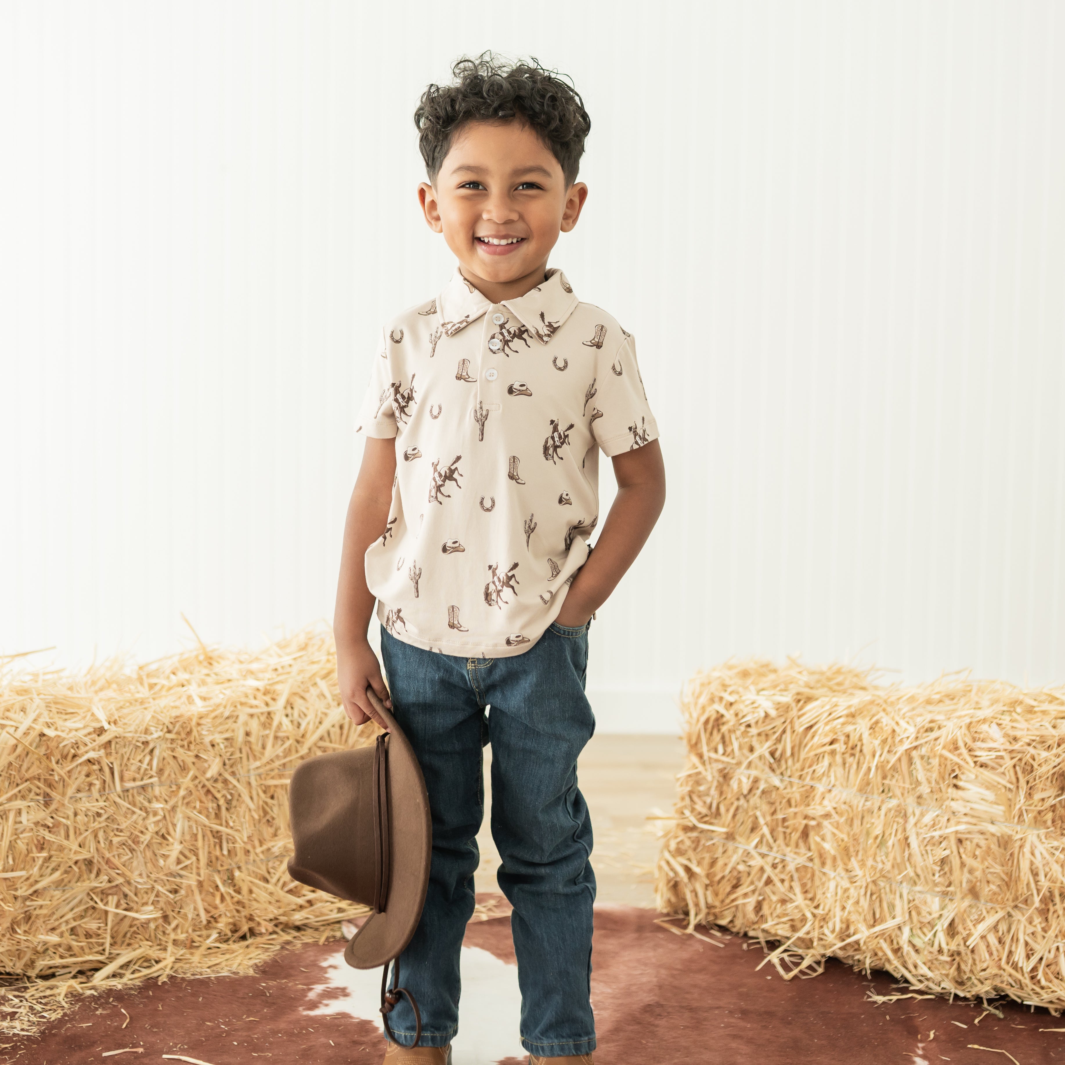 Young boy standing between two hay bales wearing the Toddler Short Sleeve Polo in Classic Cowboy paired with jeans holding a cowboy hat