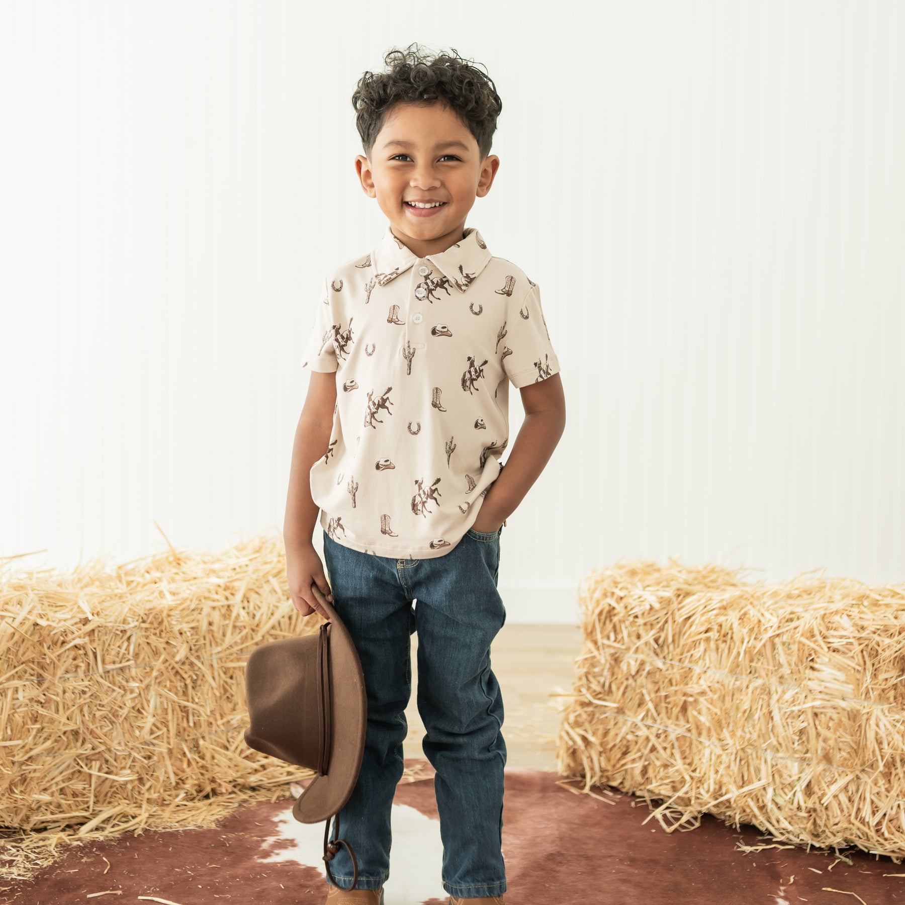 Young boy standing between two hay bales wearing the Toddler Short Sleeve Polo in Classic Cowboy paired with jeans holding a cowboy hat
