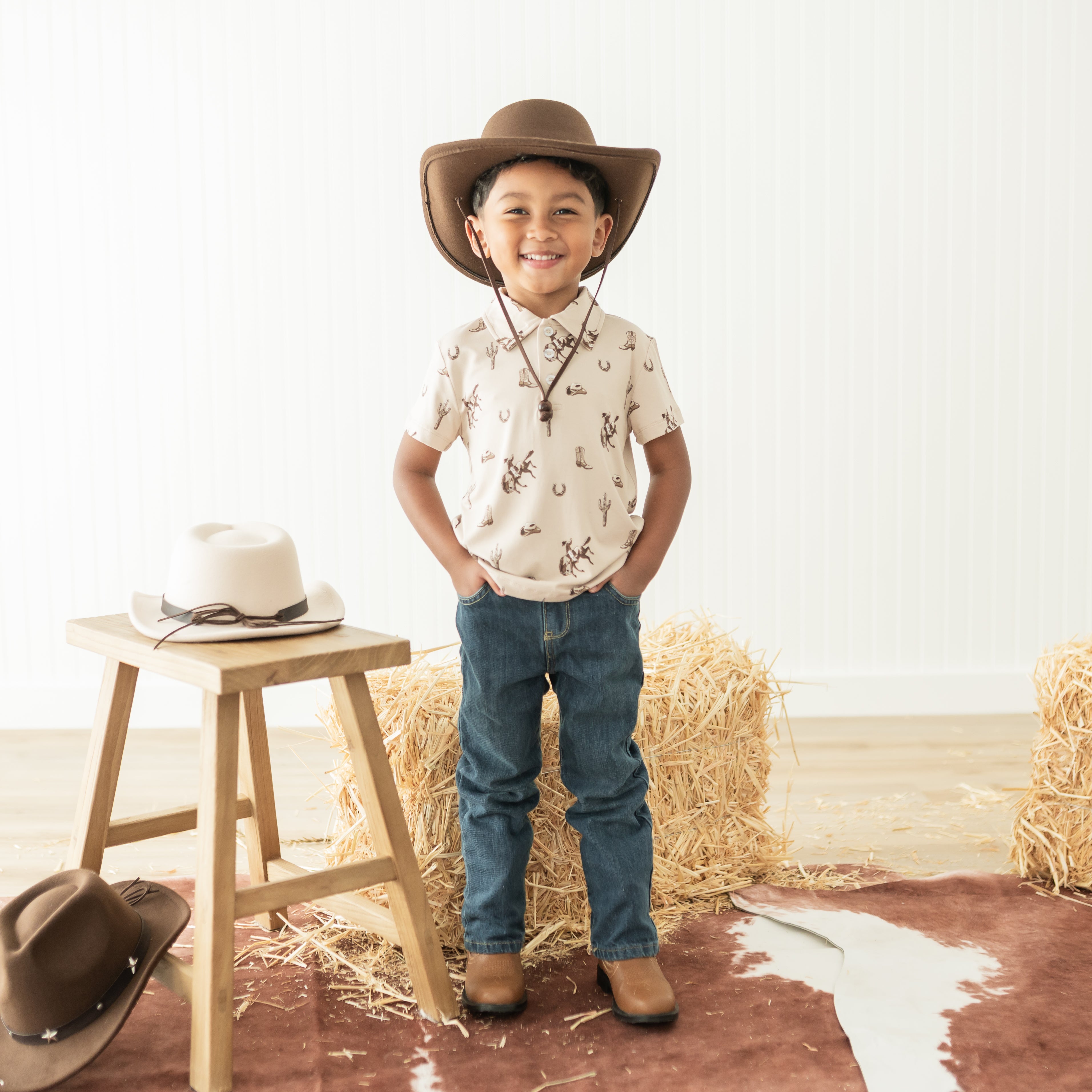 Smiling young boy standing in front of a hay bale wearing the Toddler Short Sleeve Polo in Classic Cowboy with a brown cowboy hat and medium wash jeans