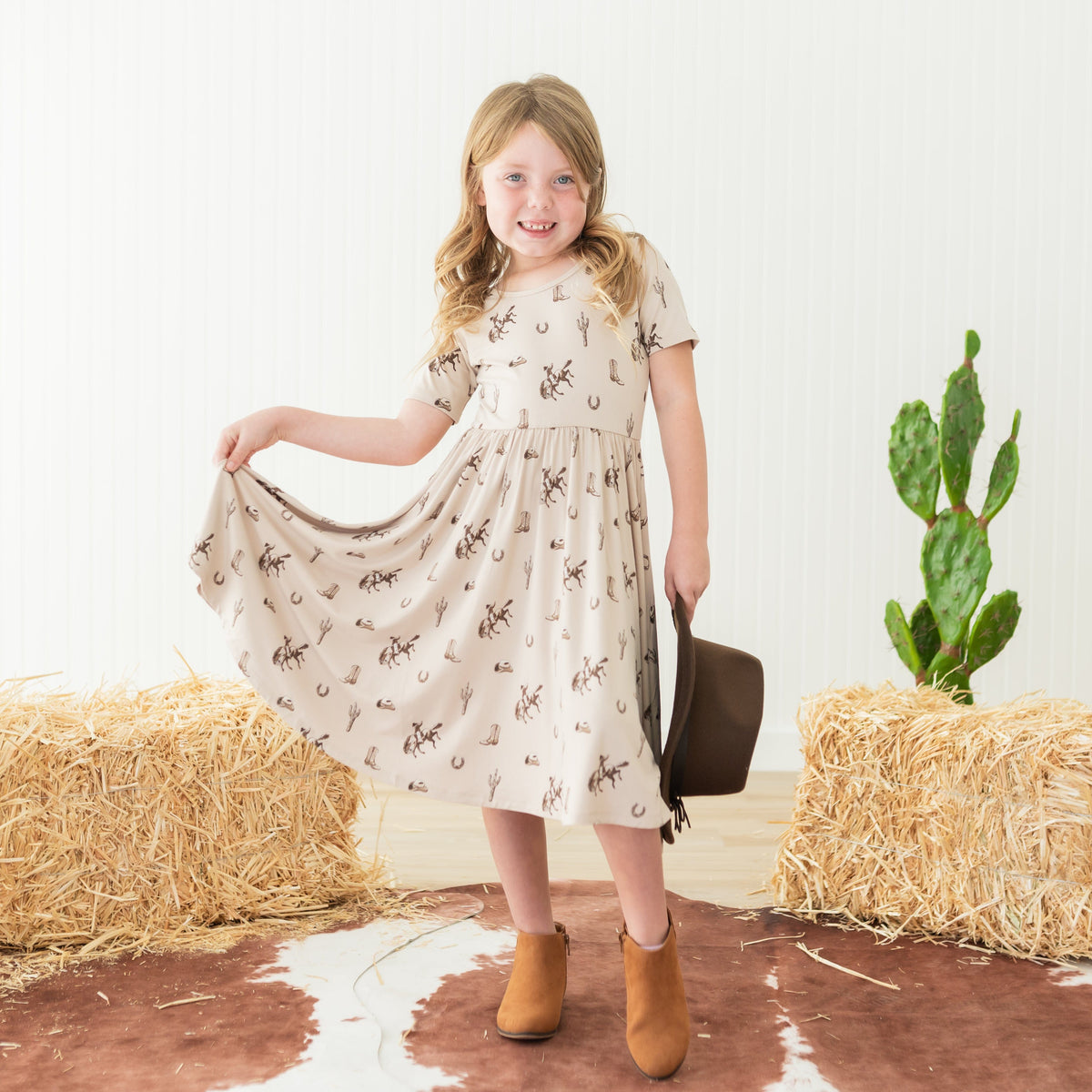Young girl standing in front of a western themed backdrop wearing the Twirl Dress in Classic Cowboy holding the bottom hem of the dress in one hand and a brown cowboy hat in the other