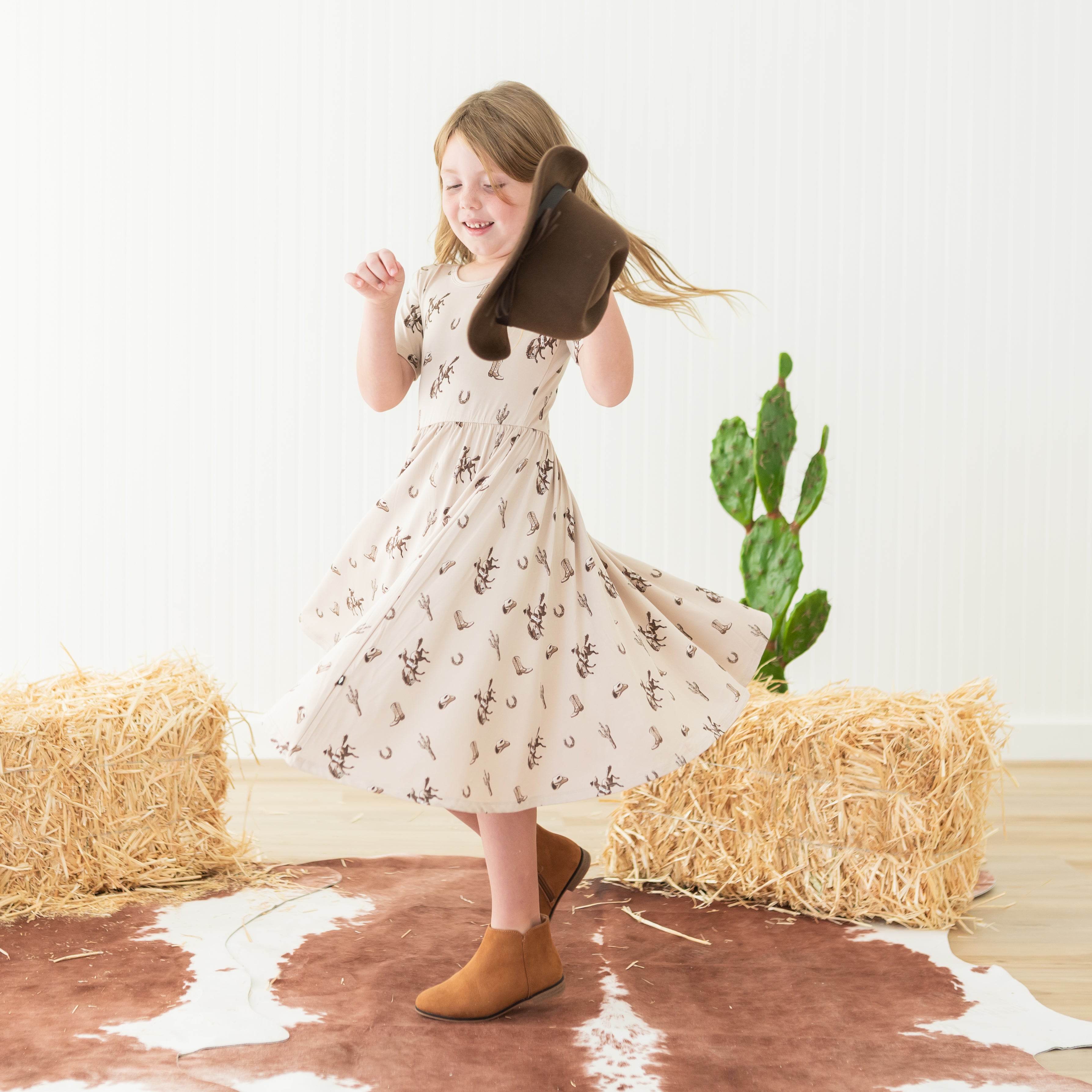 Young girl twirling wearing the Twirl Dress in Classic Cowboy holding a cowboy hat in one hand