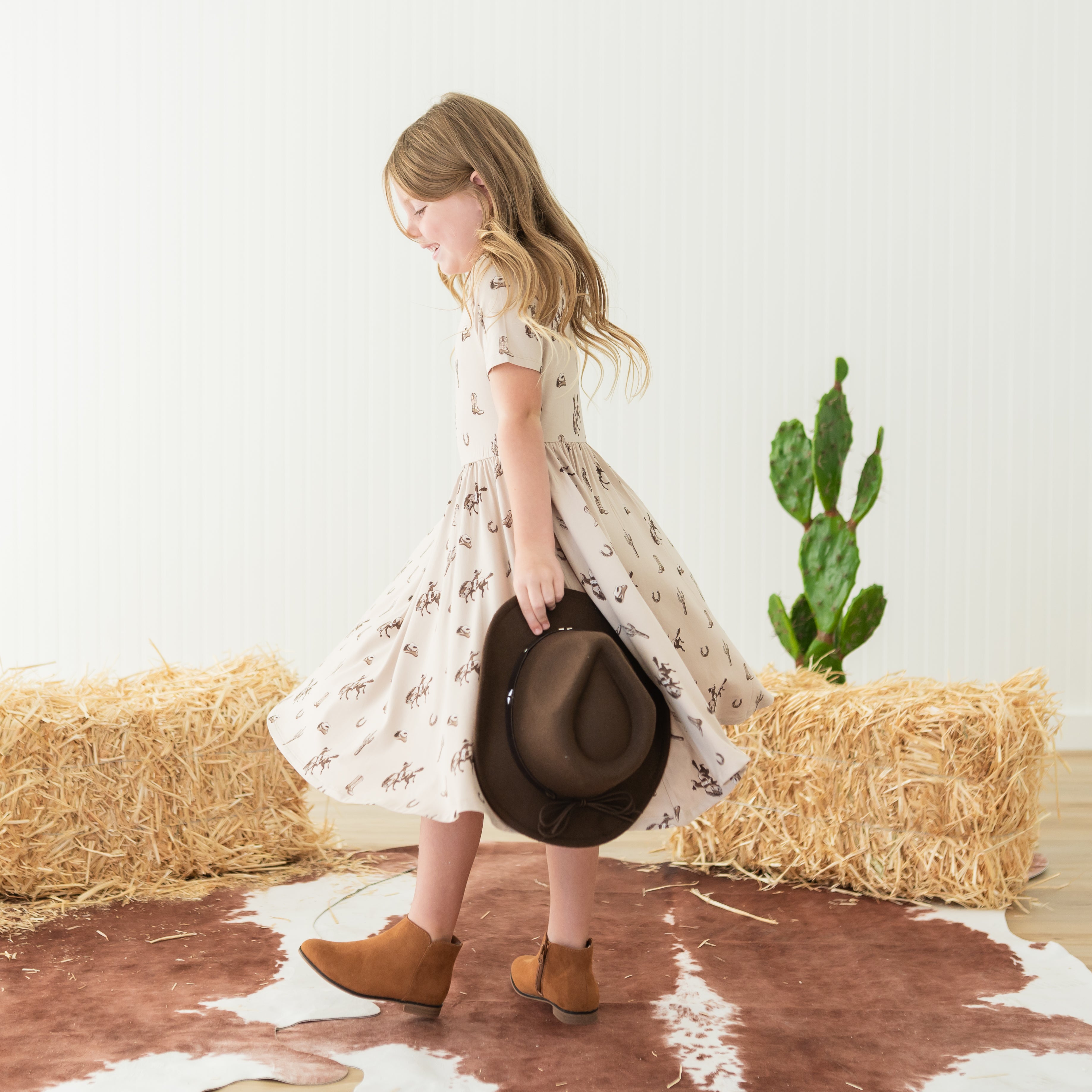 Young girl twirling in a Twirl Dress in Classic Cowboy with a cowboy hat in one hand
