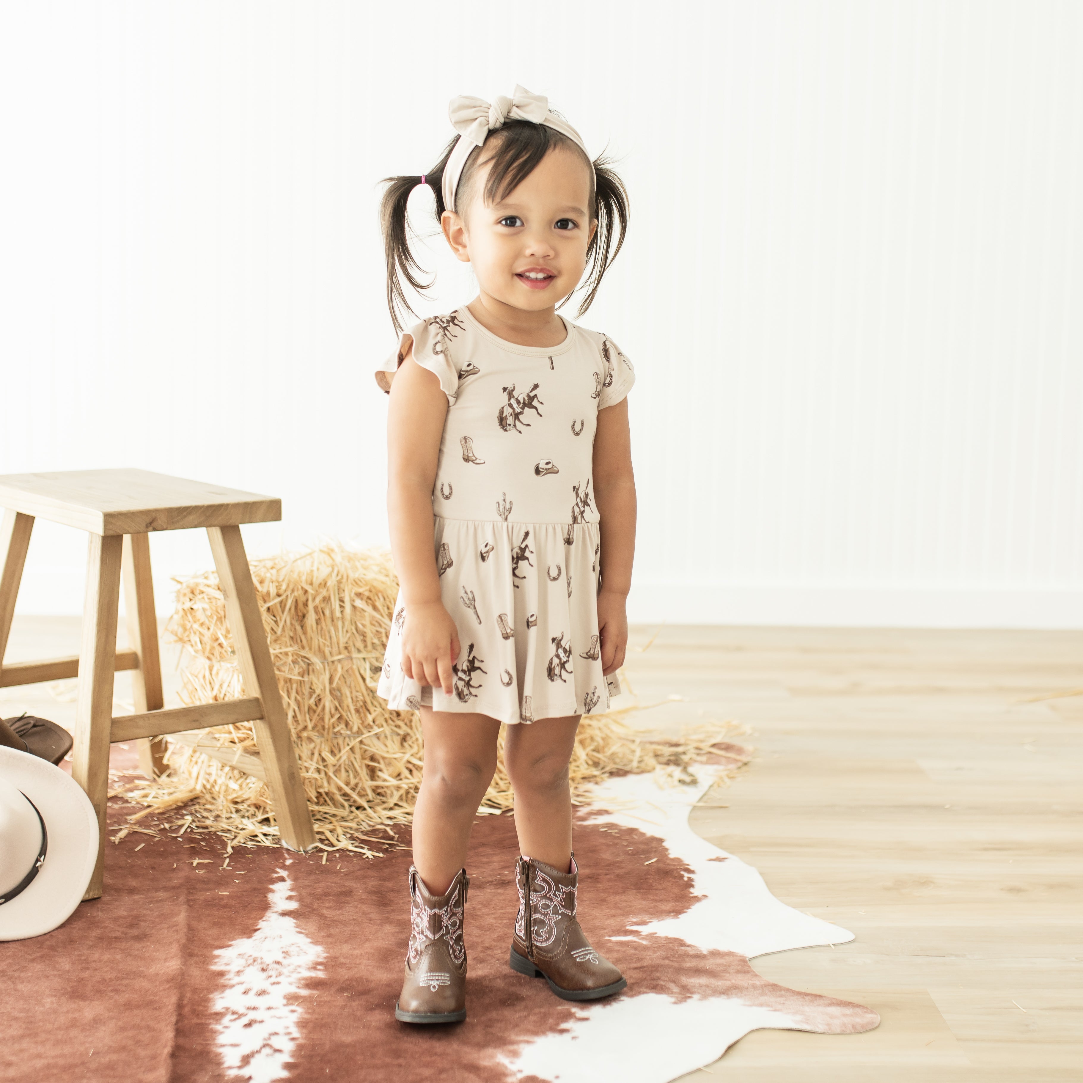 Young girl standing in front of a wooden stool and haybale wearing the Twirl Bodysuit Dress in Classic Cowboy with cowboy boots