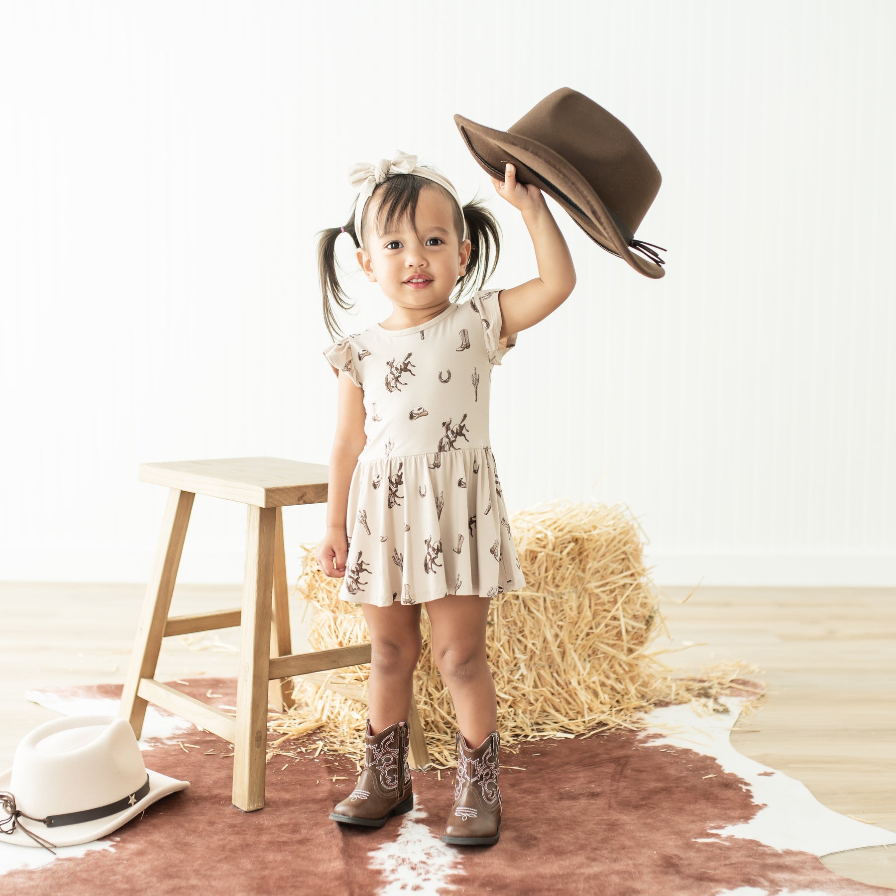 Young girl standing in front of a wooden stool and hay bale wearing the Twirl Bodysuit Dress in Classic Cowboy holding a brown cowboy hat