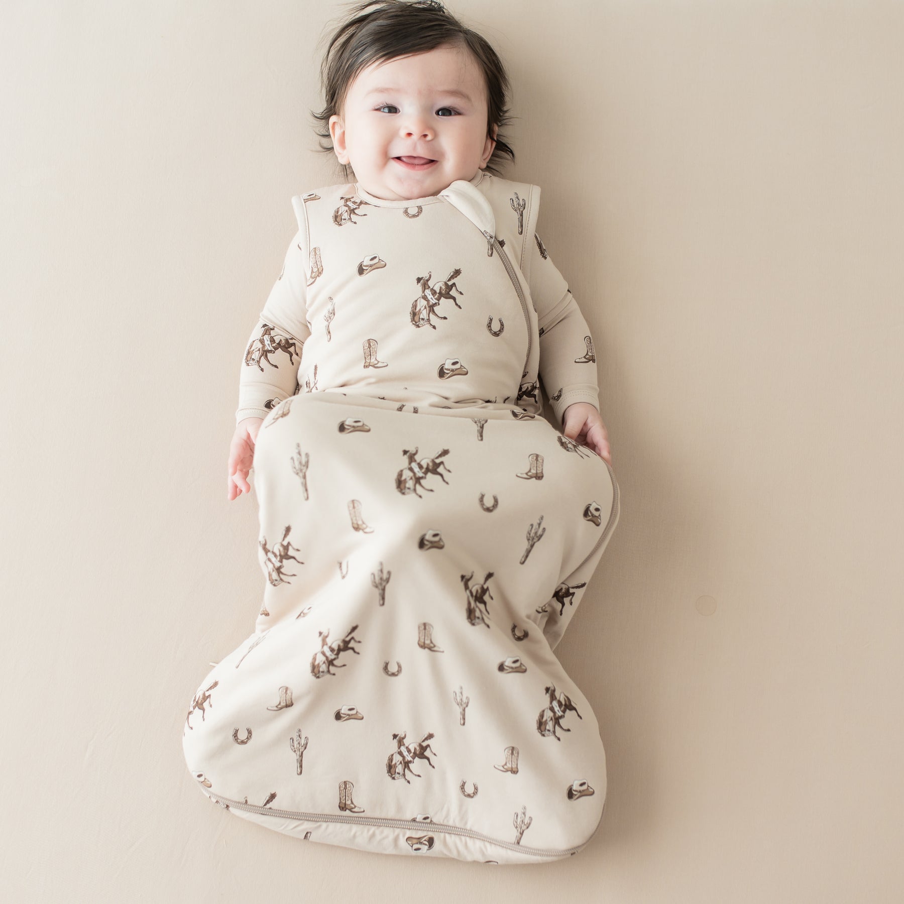 Smiling infant laying on a light beige surface wearing the Sleep Bag in Classic Cowboy 1.0 with their legs bent and in the air