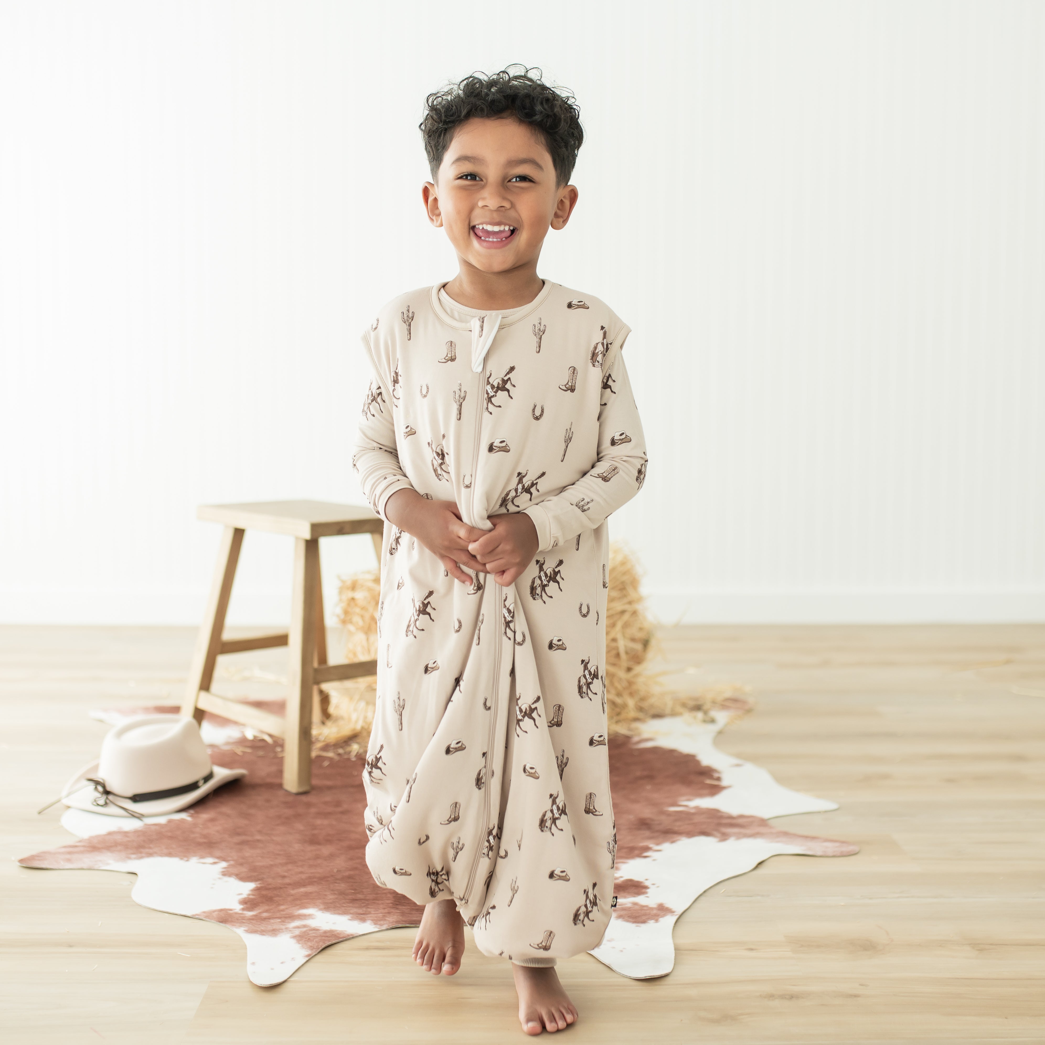 Smiling boy walking wearing the Sleep Bag Walker in Classic Cowboy 1.0 in front of a western inspired backdrop